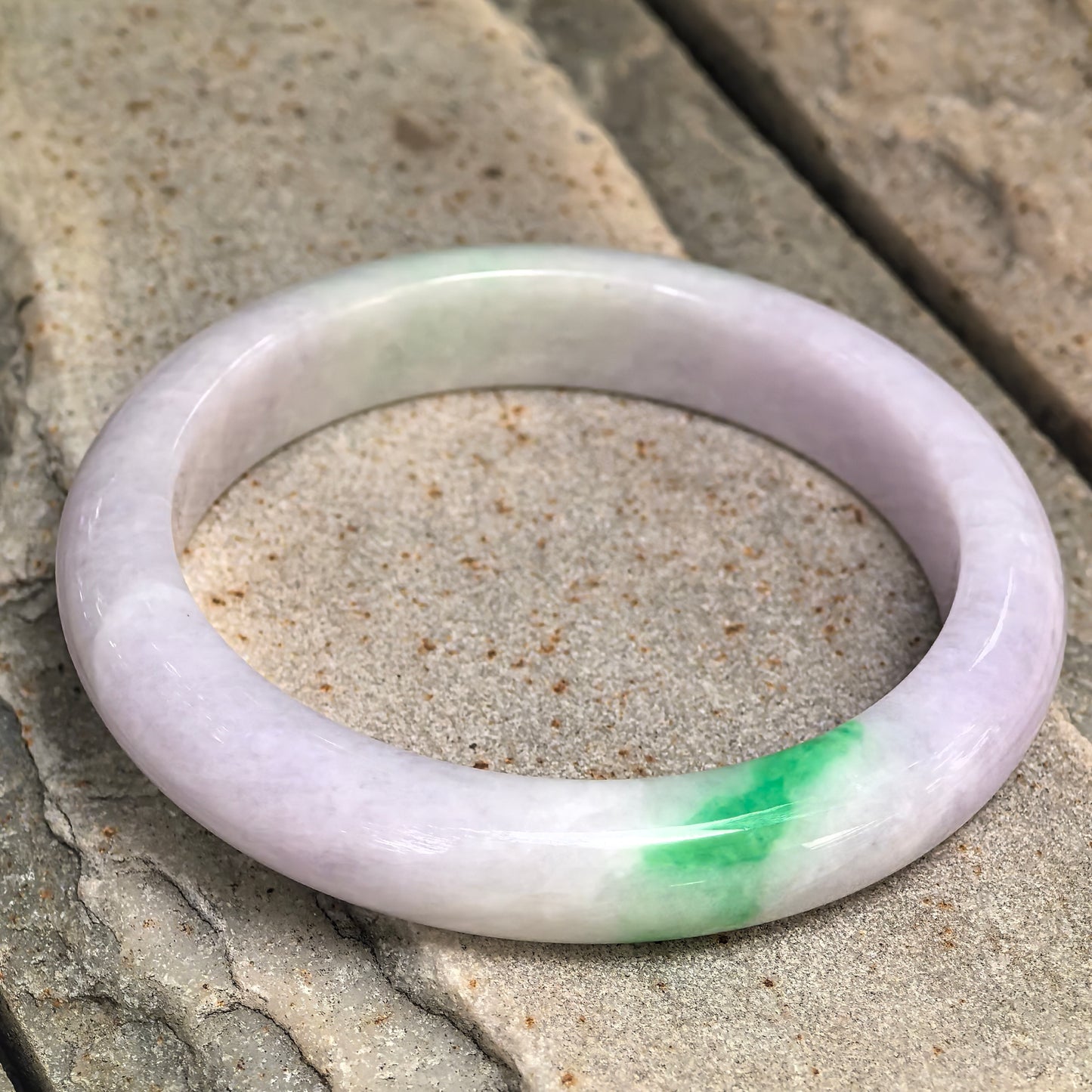 Natural Grade-A pale lavender and bright green jadeite D-shape bangle displayed on stone, showing the contrasting color section.