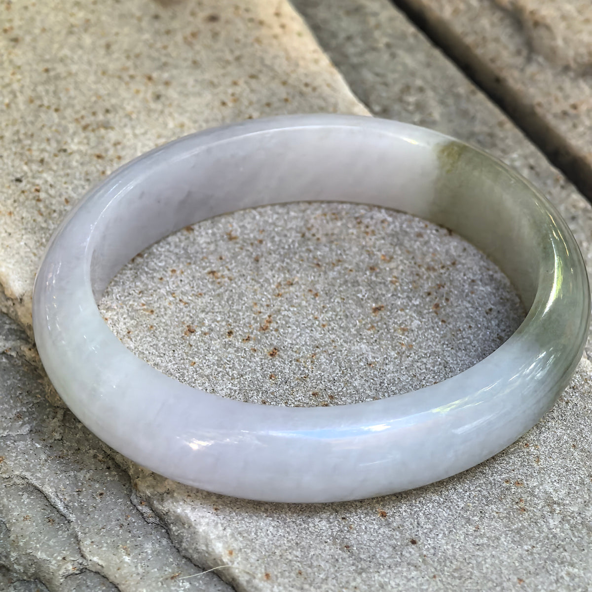 Natural Grade-A glutinous pale lavender and oil green oval jadeite bangle displayed on stone, showing its broad polished profile.
