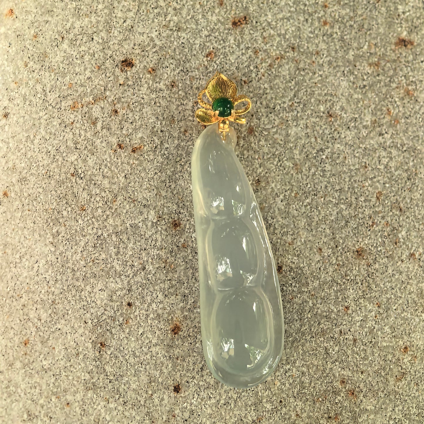 Natural light green jadeite bean pod pendant displayed on stone with 18K gold floral bail and green accent stone.