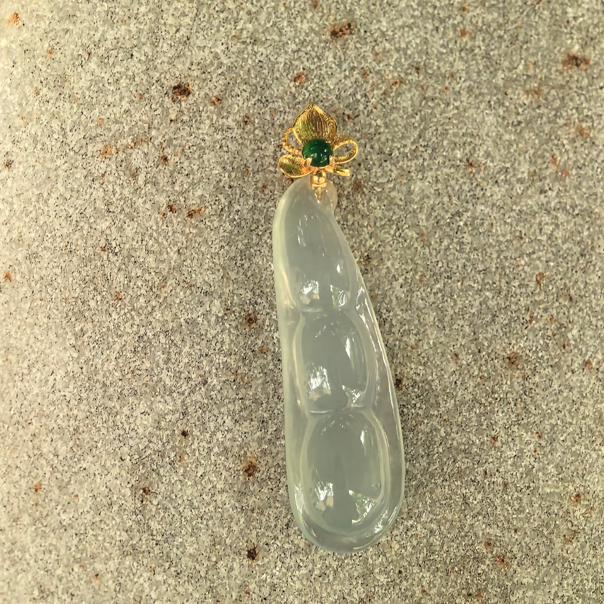 Natural light green jadeite bean pod pendant displayed on stone with 18K gold floral bail and green accent stone.