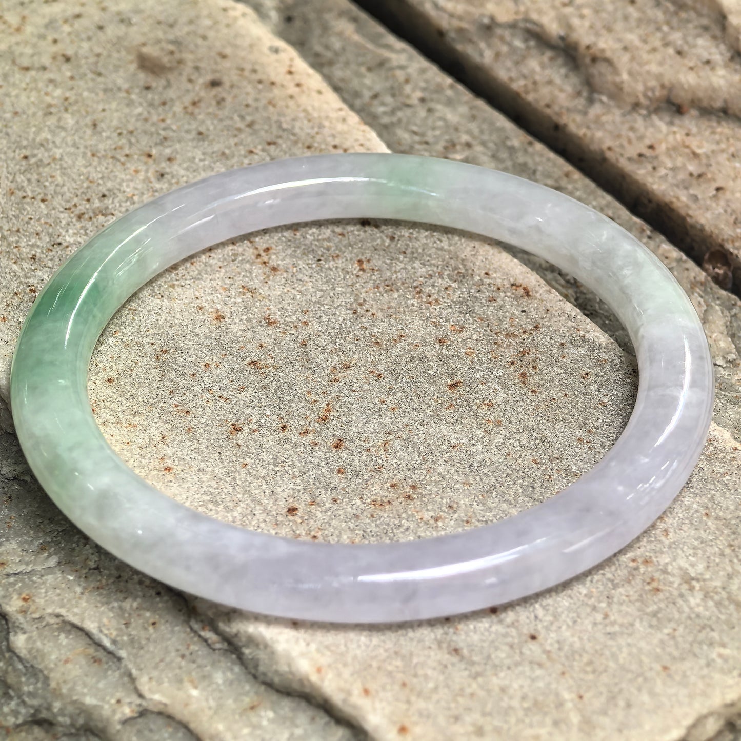 Natural Grade-A icy glutinous lavender-green jadeite bangle displayed on stone, showing its slender round shape and polished luster.