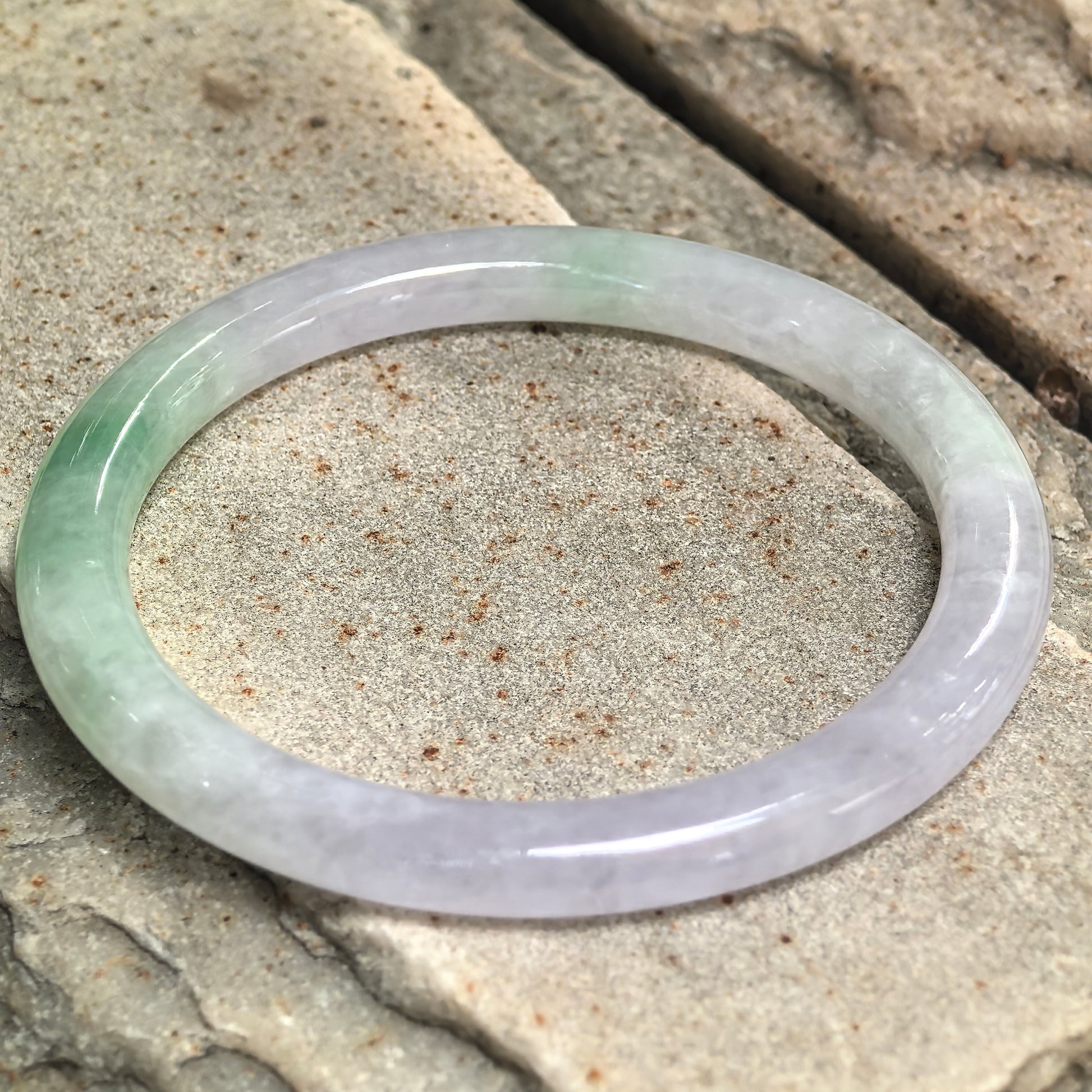 Natural Grade-A icy glutinous lavender-green jadeite bangle displayed on stone, showing its slender round shape and polished luster.