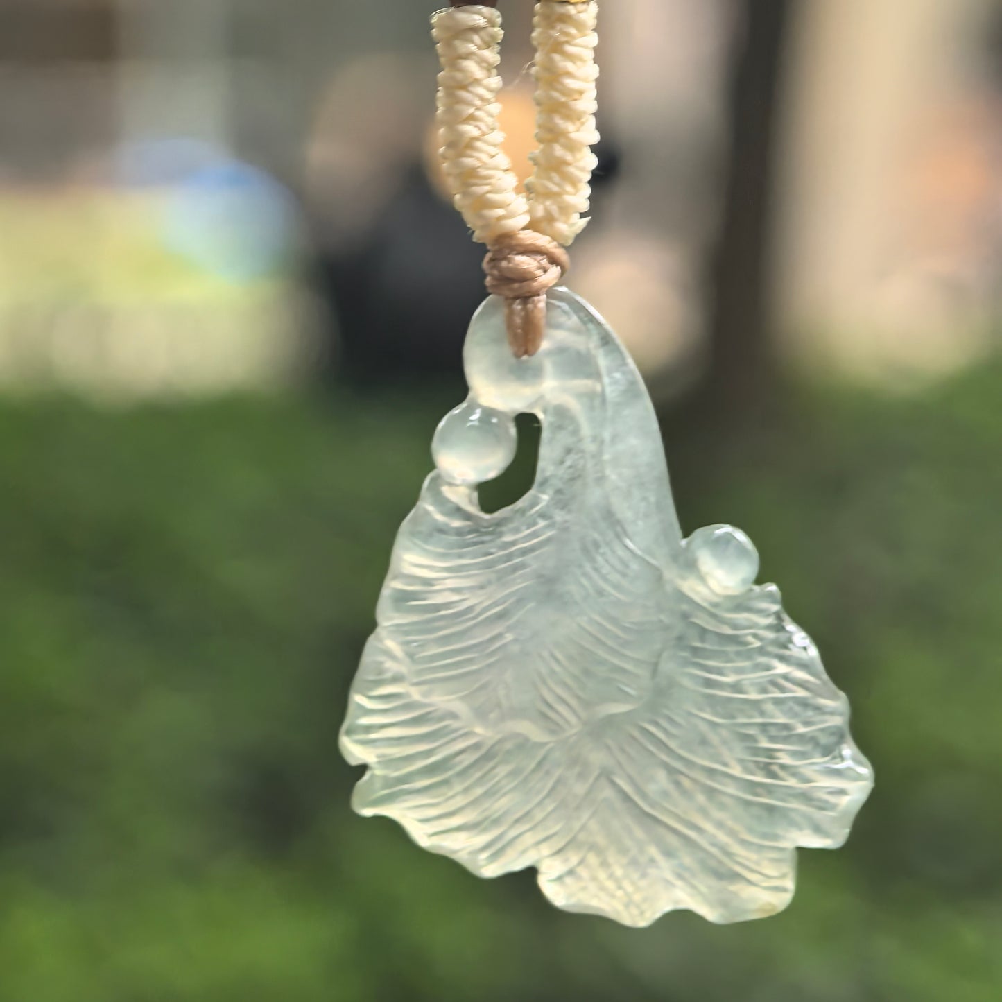 Icy light green ginkgo leaf jadeite pendant hanging outdoors, showing its translucent carved leaf design.