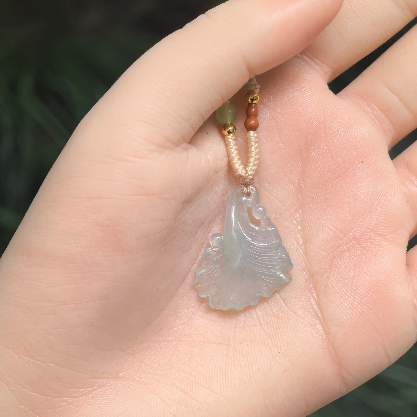 Natural Grade-A icy light green ginkgo leaf jadeite pendant shown in hand with a woven cord for scale.