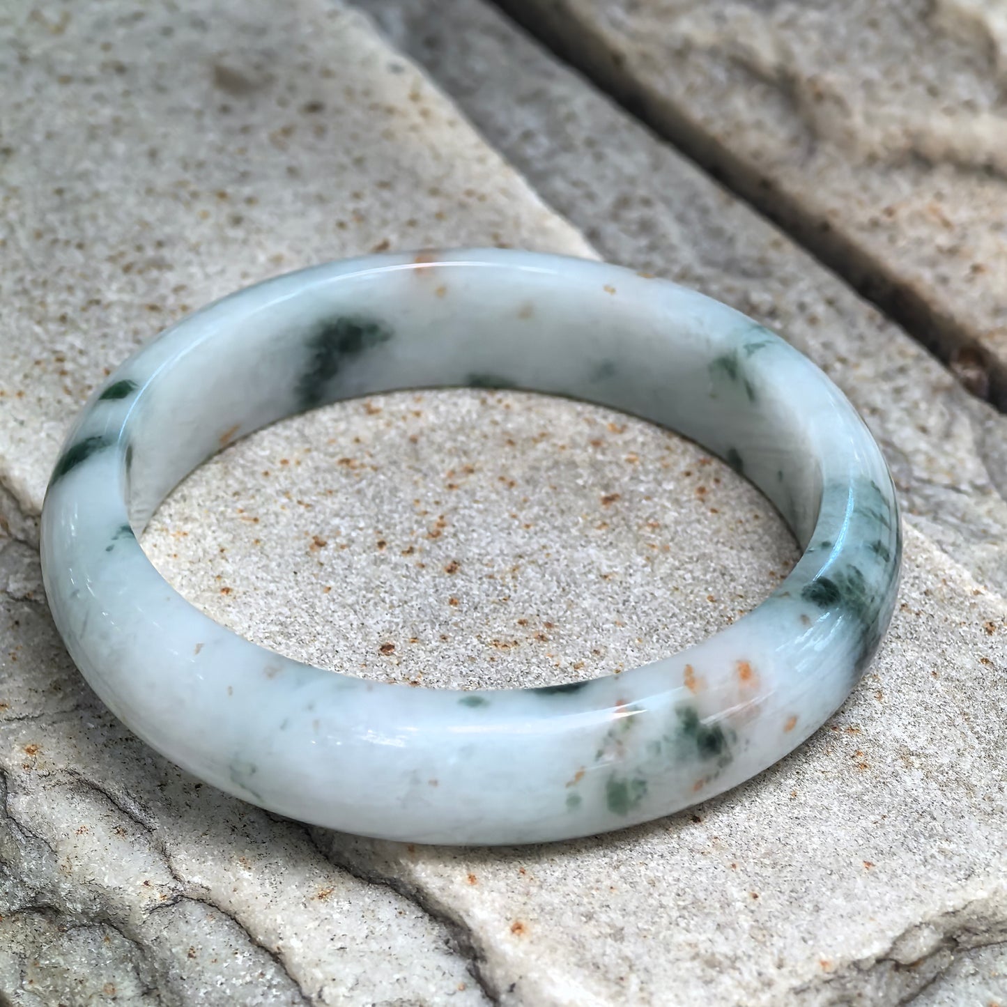 Natural Grade-A fine glutinous jadeite D-shape bangle displayed on stone showing milk white base, green floating patterns, and natural golden flecks.