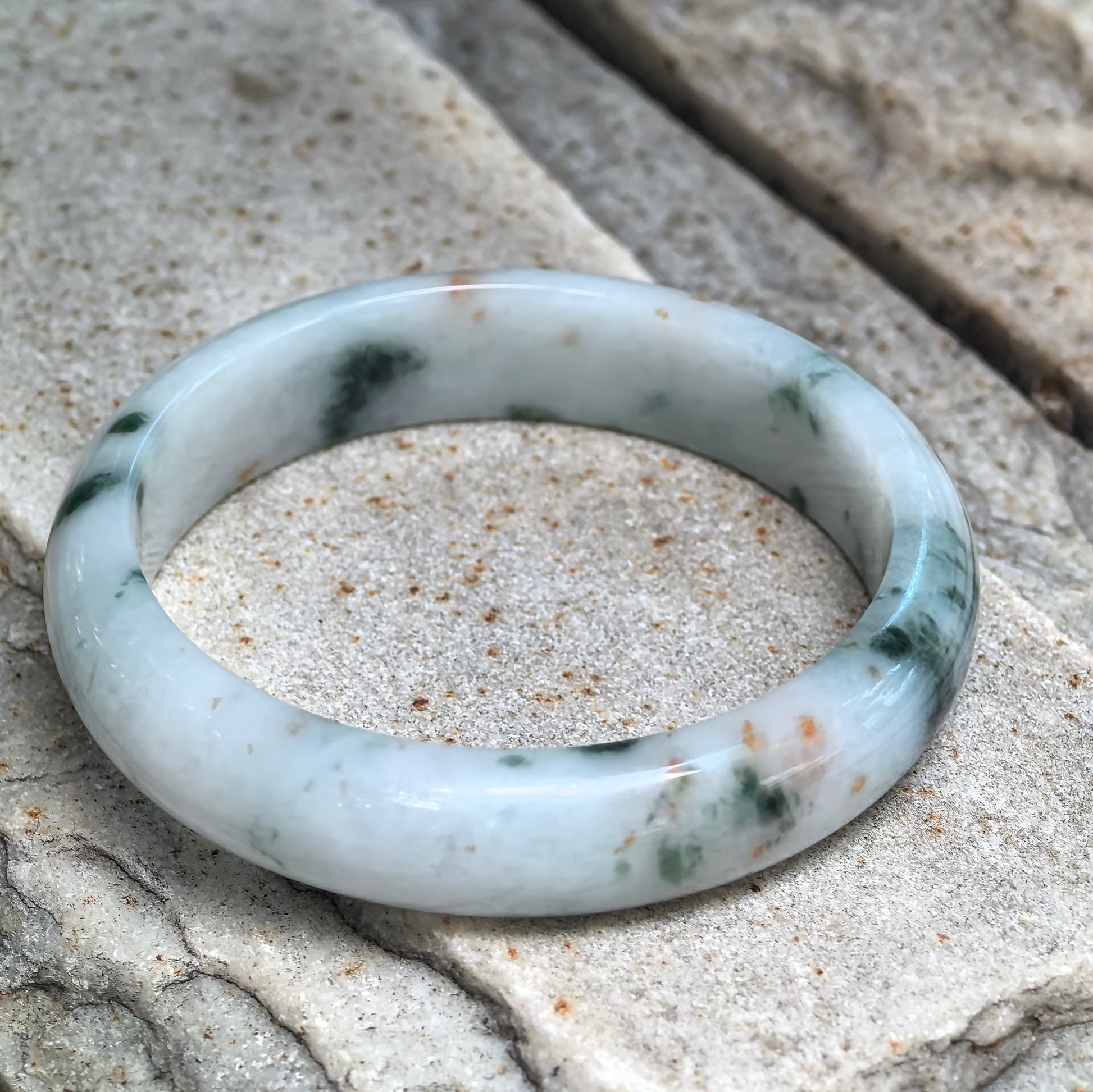 Natural Grade-A fine glutinous jadeite D-shape bangle displayed on stone showing milk white base, green floating patterns, and natural golden flecks.