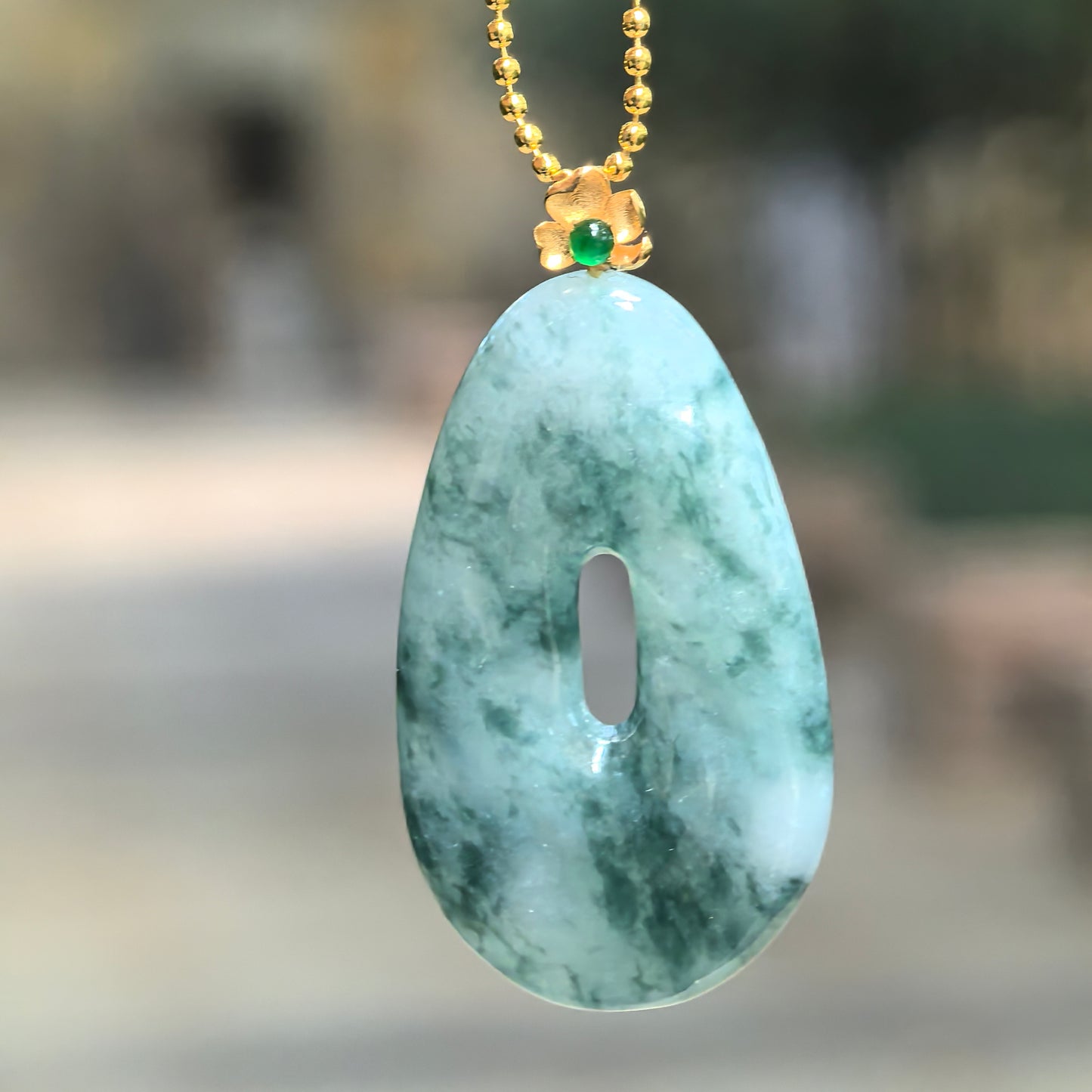 Hanging view of Natural Grade-A icy glutinous jadeite pendant under bright light showing lighter green translucency, mottled color, and 18K gold floral bail.