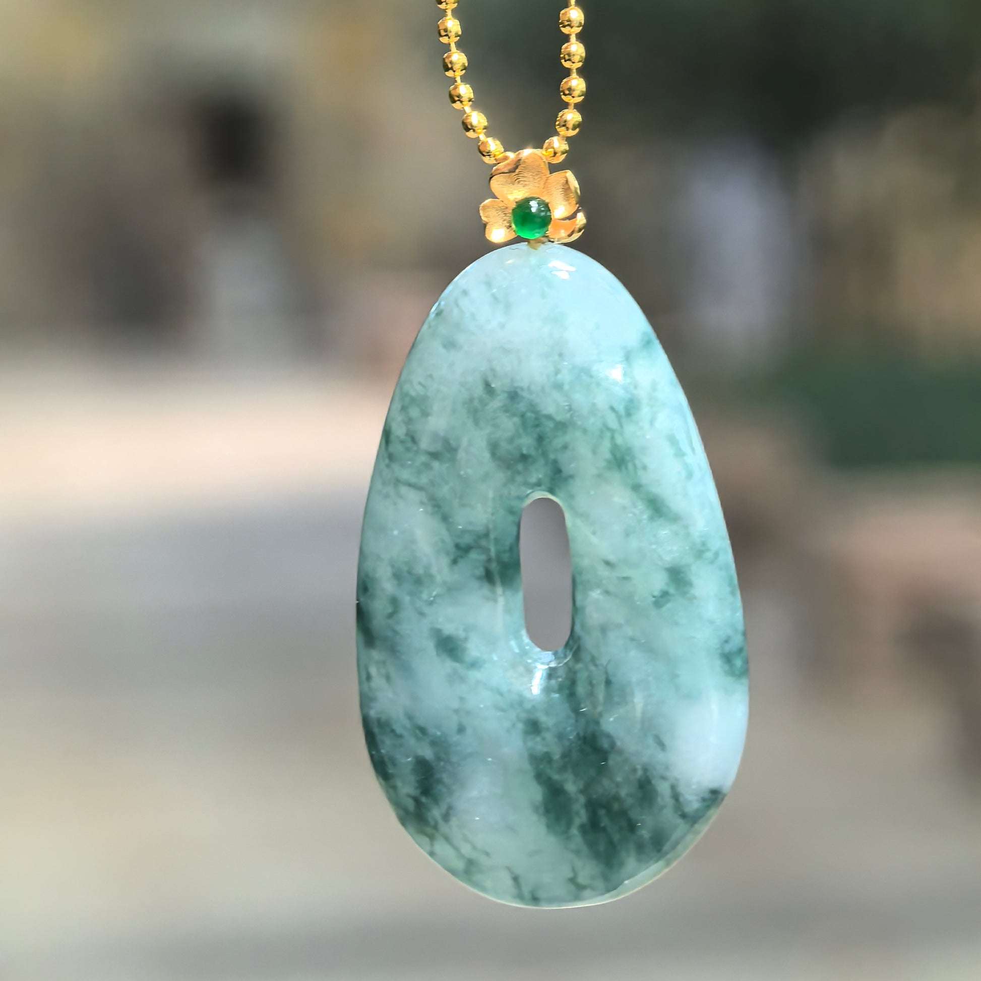 Hanging view of Natural Grade-A icy glutinous jadeite pendant under bright light showing lighter green translucency, mottled color, and 18K gold floral bail.