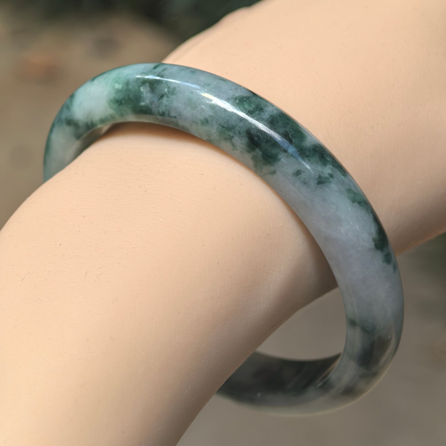Wrist view of glutinous Burmese jadeite bangle showing deep ink-green floating patterns over a light green and gray-green base.