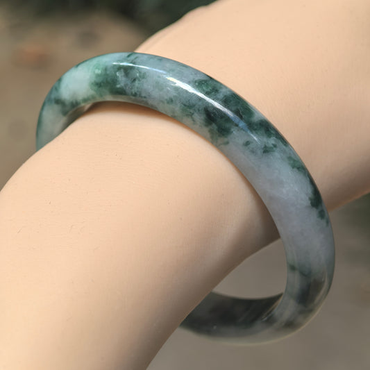 Wrist view of glutinous Burmese jadeite bangle showing deep ink-green floating patterns over a light green and gray-green base.