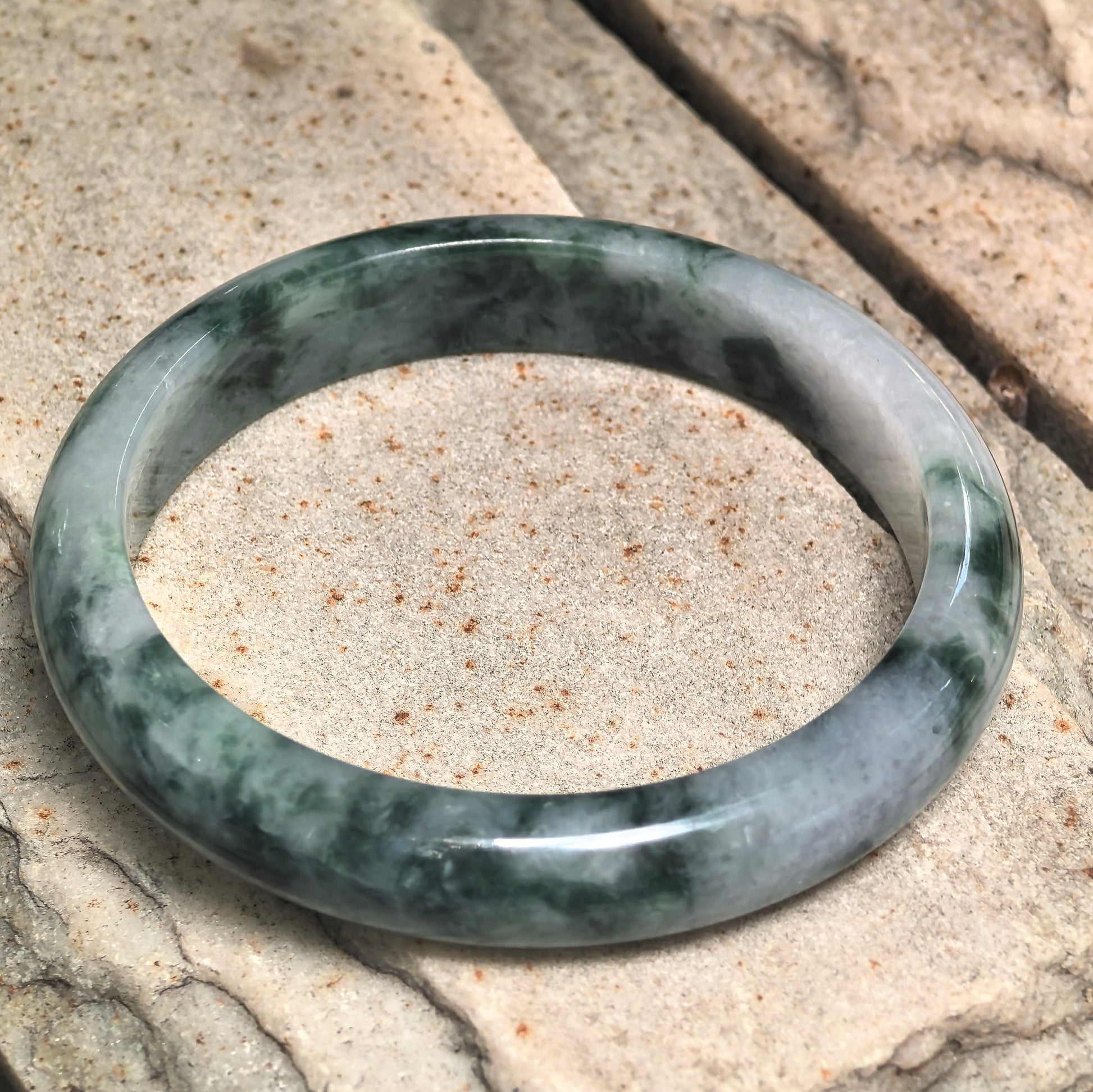 Angled view of Natural Grade-A glutinous dark green floating flower jadeite bangle on stone showing its wide D-shape profile.