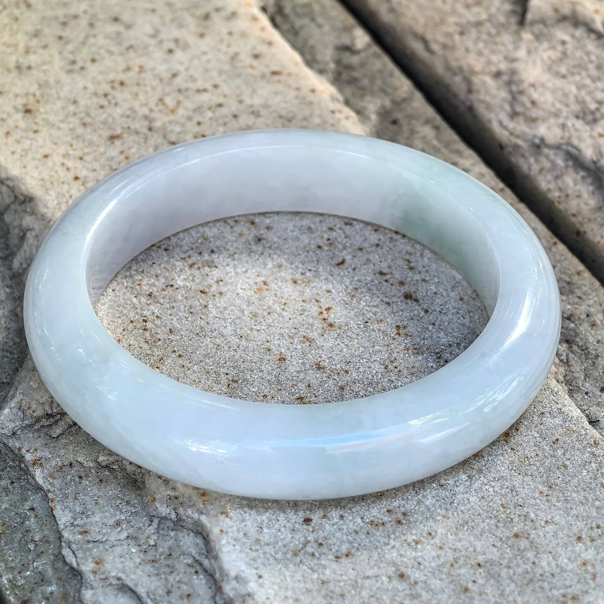 Glutinous white and light green Burmese jadeite D-shape bangle displayed at an angle on a stone surface.