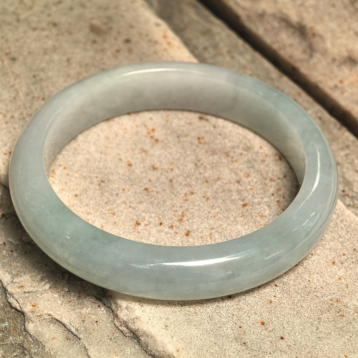 Light green jadeite bangle resting at an angle on textured stone, showing its flat inner profile and rounded polished exterior.