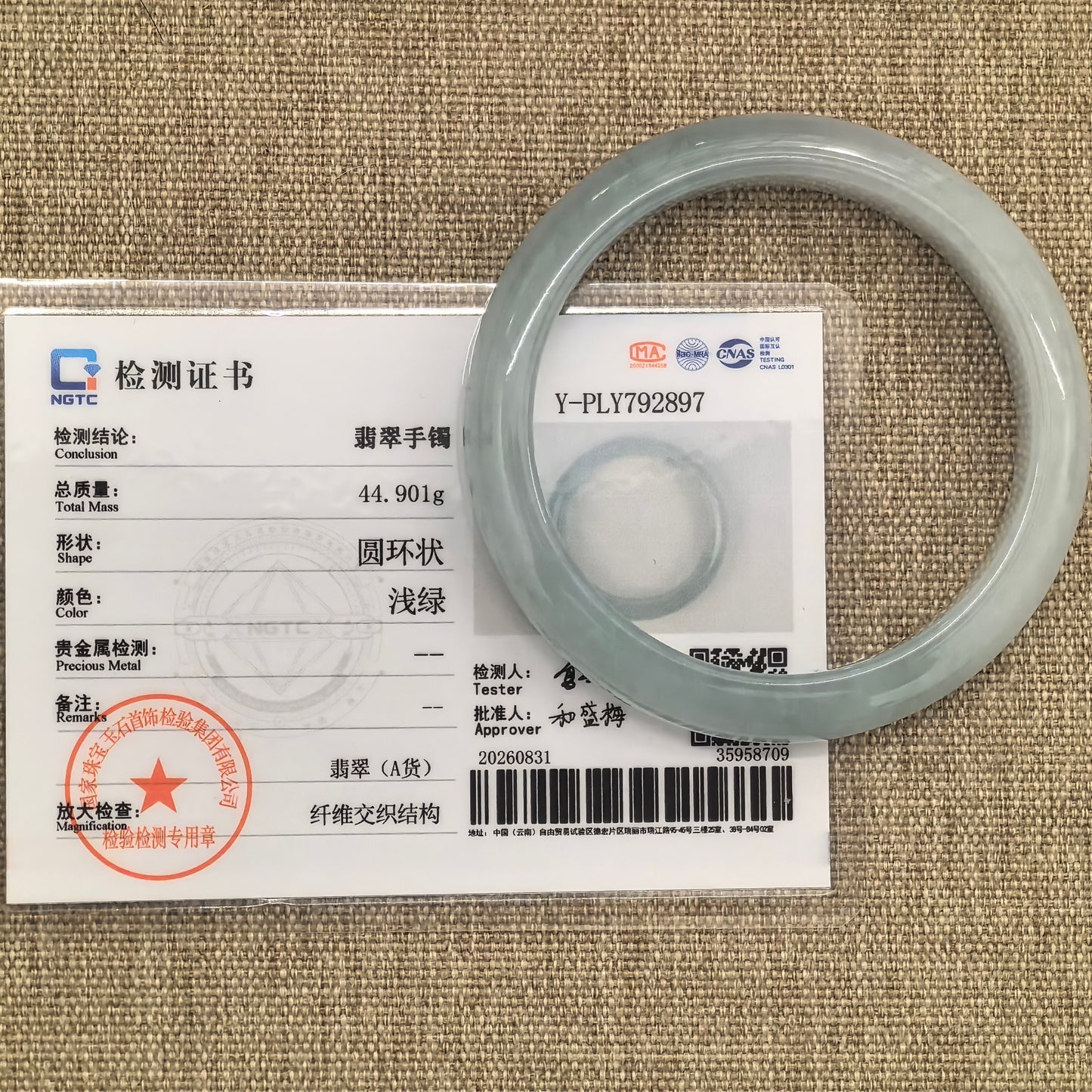 Pale green jadeite bangle displayed with its matching NGTC certificate on woven fabric, with Grade-A identification and weight visible.
