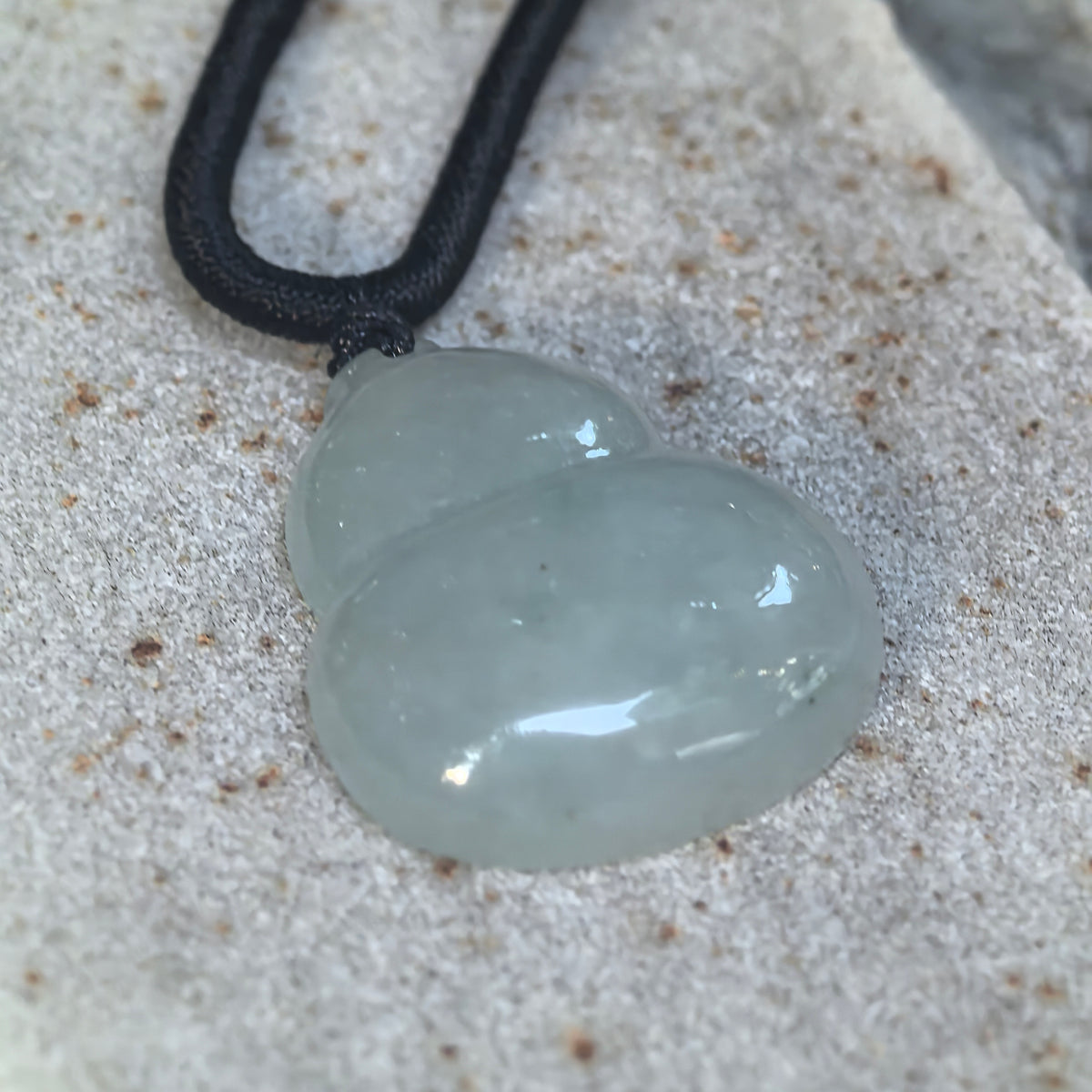 Angled view of a pale green jadeite gourd pendant on textured stone, highlighting its rounded sections and polished finish.