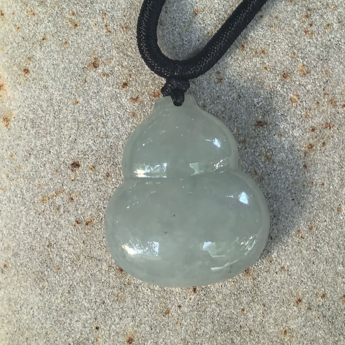 Front view of a light green jadeite gourd pendant on stone, showing the defined waist, black cord attachment, and tiny dark specks on the lower section.
