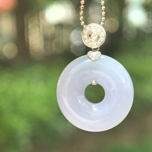 Lavender jadeite donut pendant with an 18K gold and diamond setting, hanging from a chain against a blurred green background.
