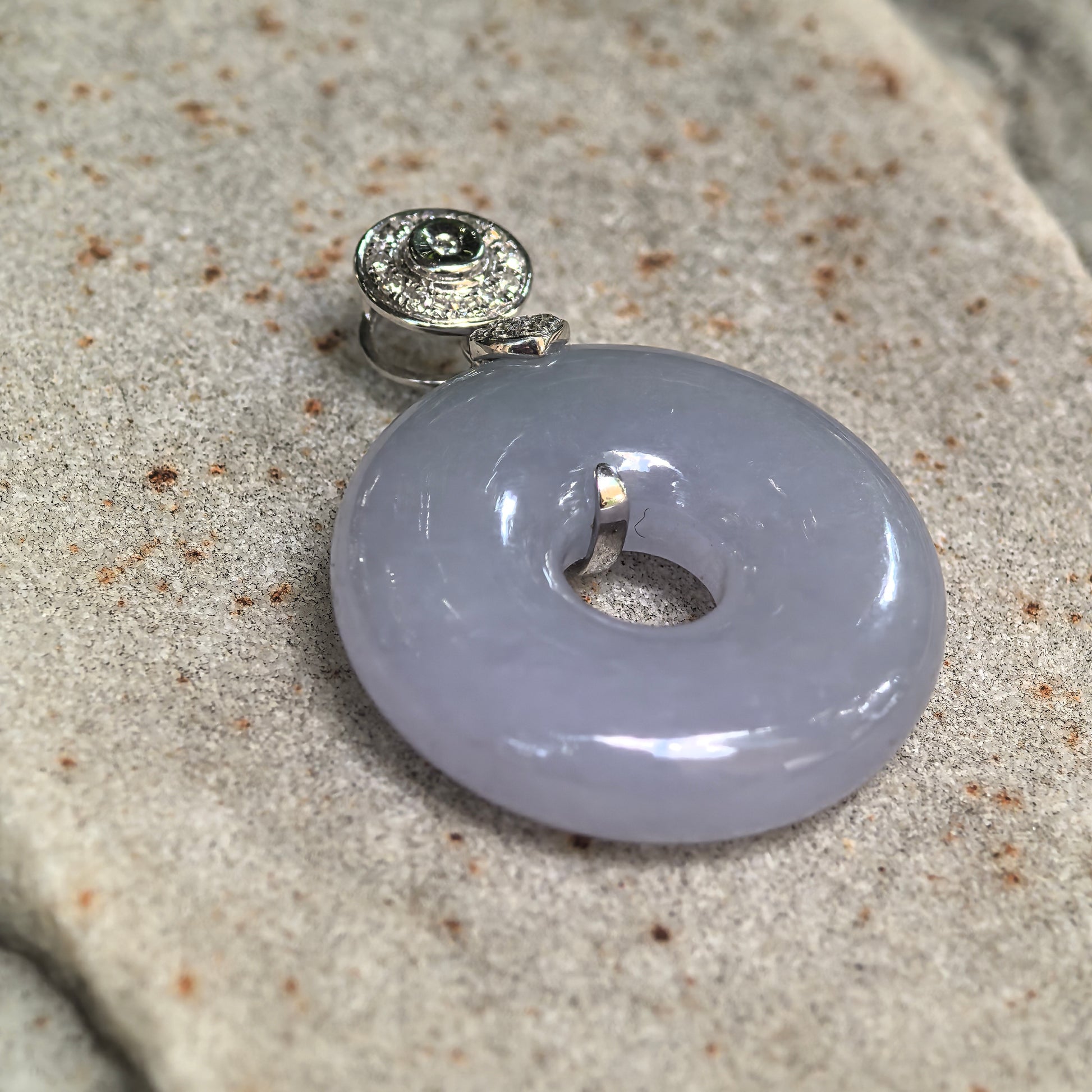 Angled close-up of a lavender jadeite donut pendant on textured stone, showing its rounded profile and raised diamond-set bail.