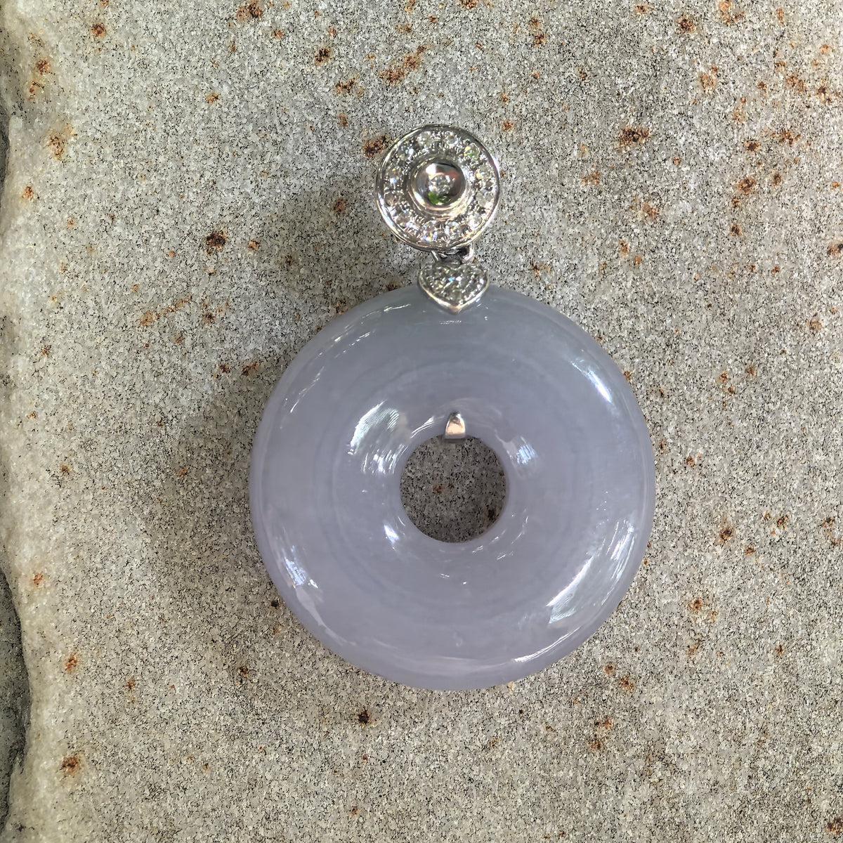 Front view of a lavender jadeite donut pendant on pale stone, with a circular diamond-set bail and small heart-shaped connector.