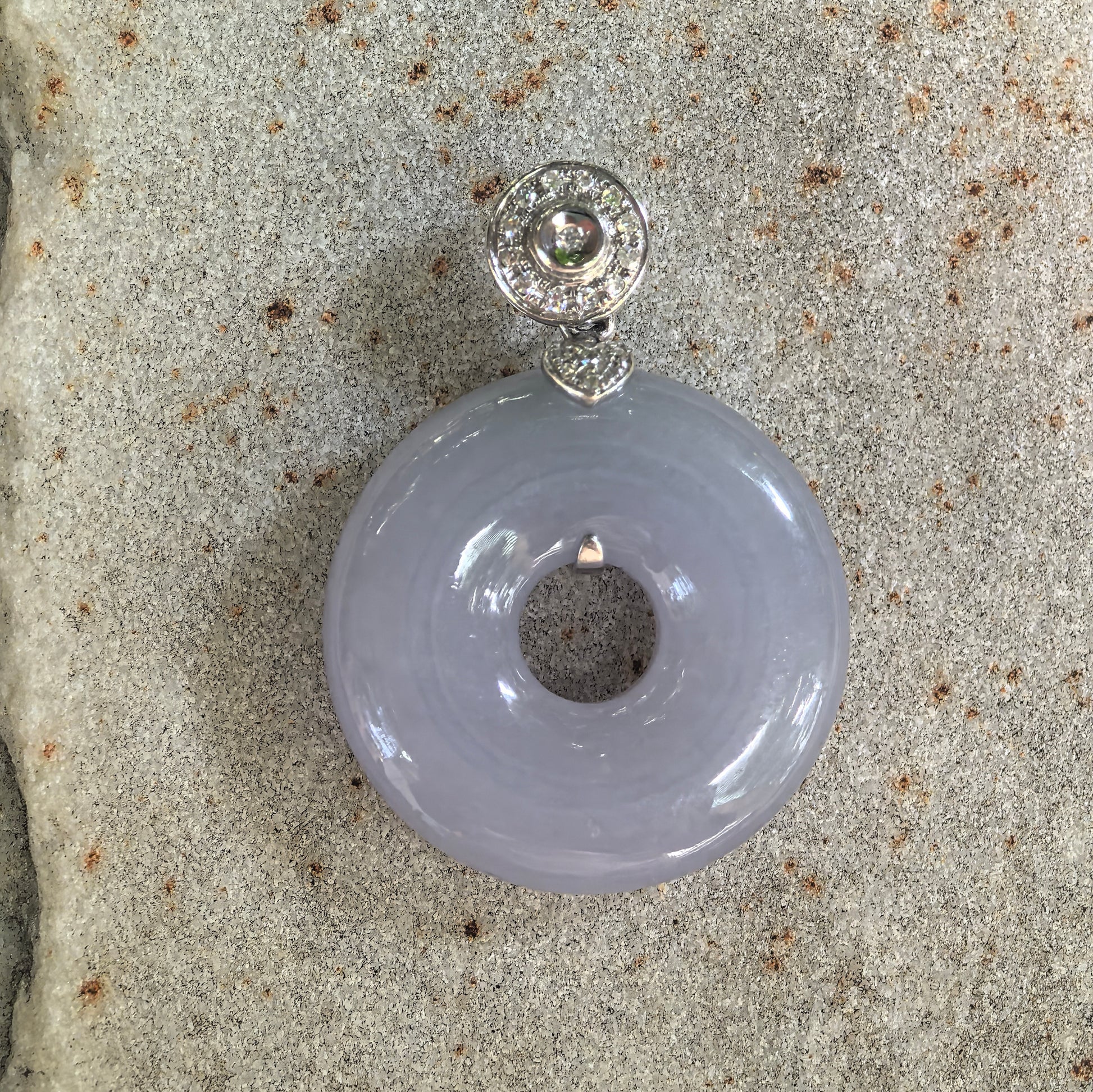 Front view of a lavender jadeite donut pendant on pale stone, with a circular diamond-set bail and small heart-shaped connector.