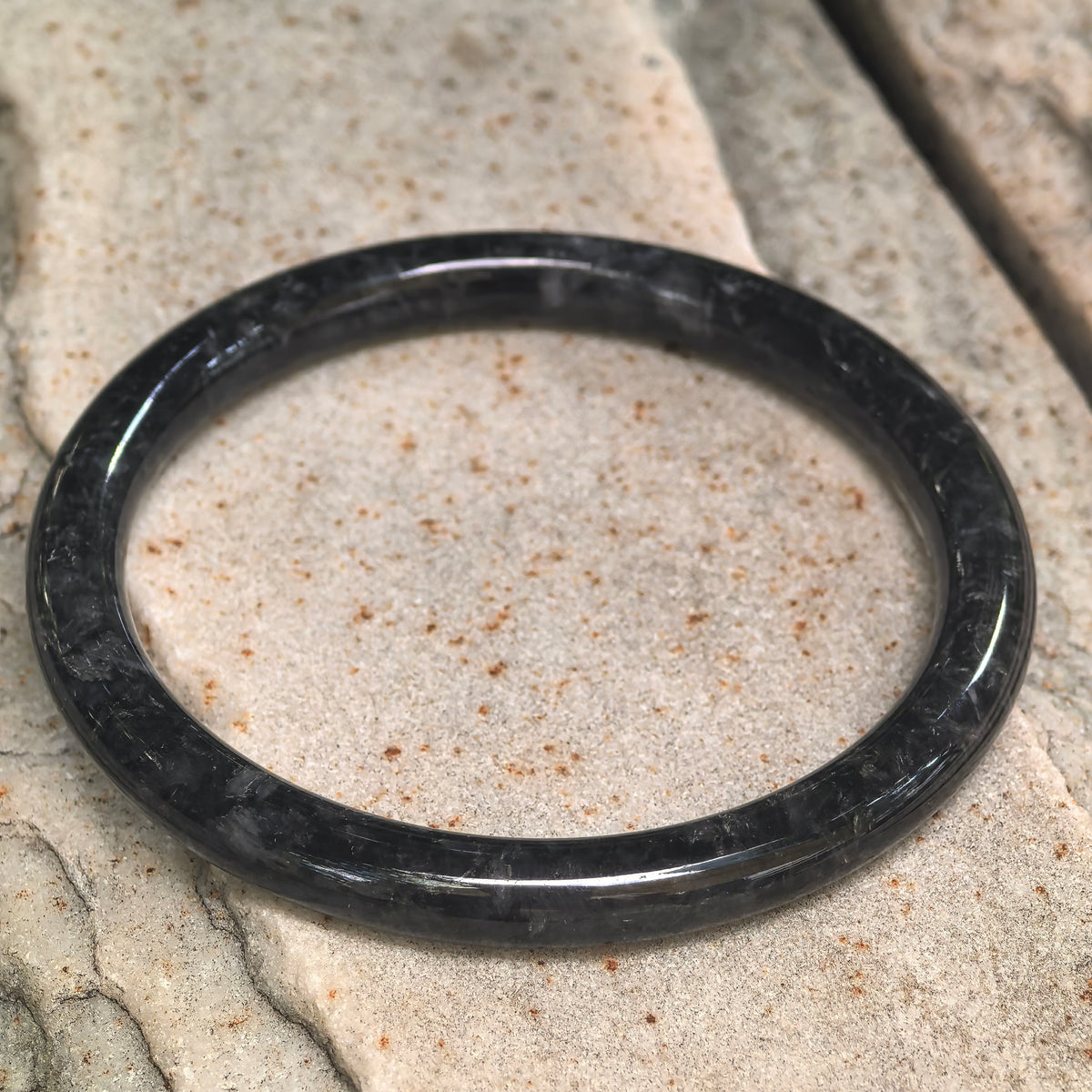 Black and gray round-bar jadeite bangle resting at an angle on light textured stone.
