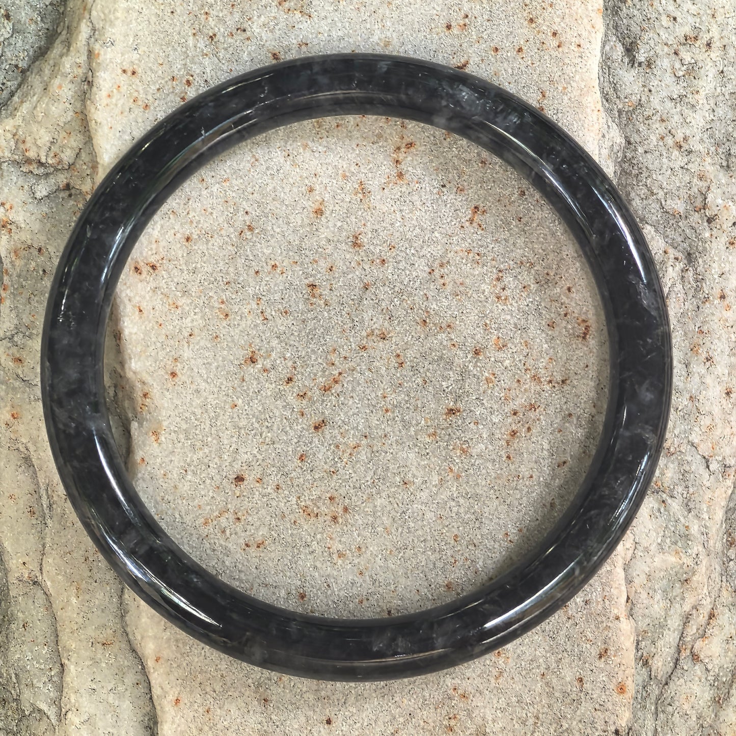 Top-down view of a slim circular jadeite bangle with smoky gray patterns against a light stone background.