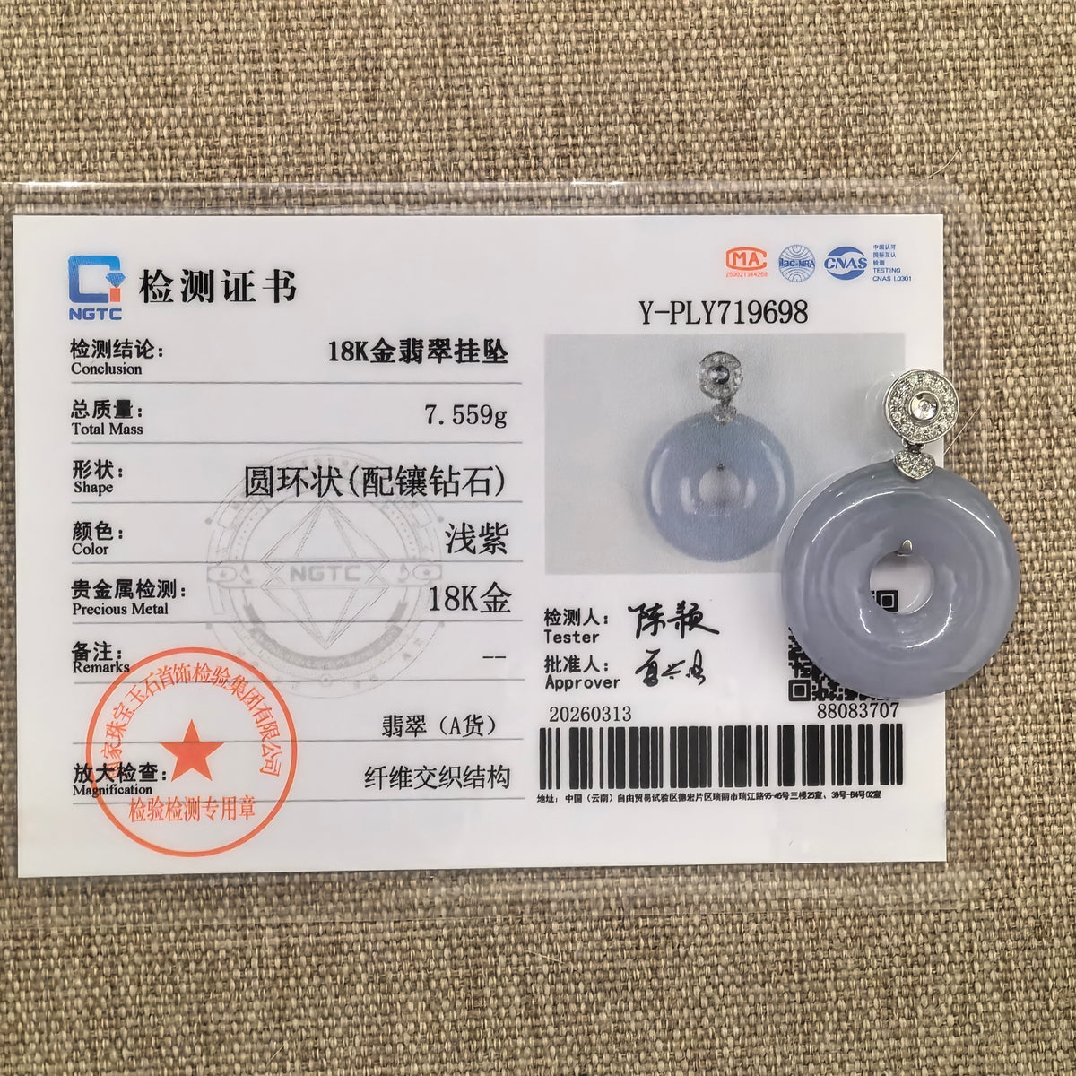 Lavender jadeite donut pendant displayed beside its matching NGTC certificate on tan woven fabric.