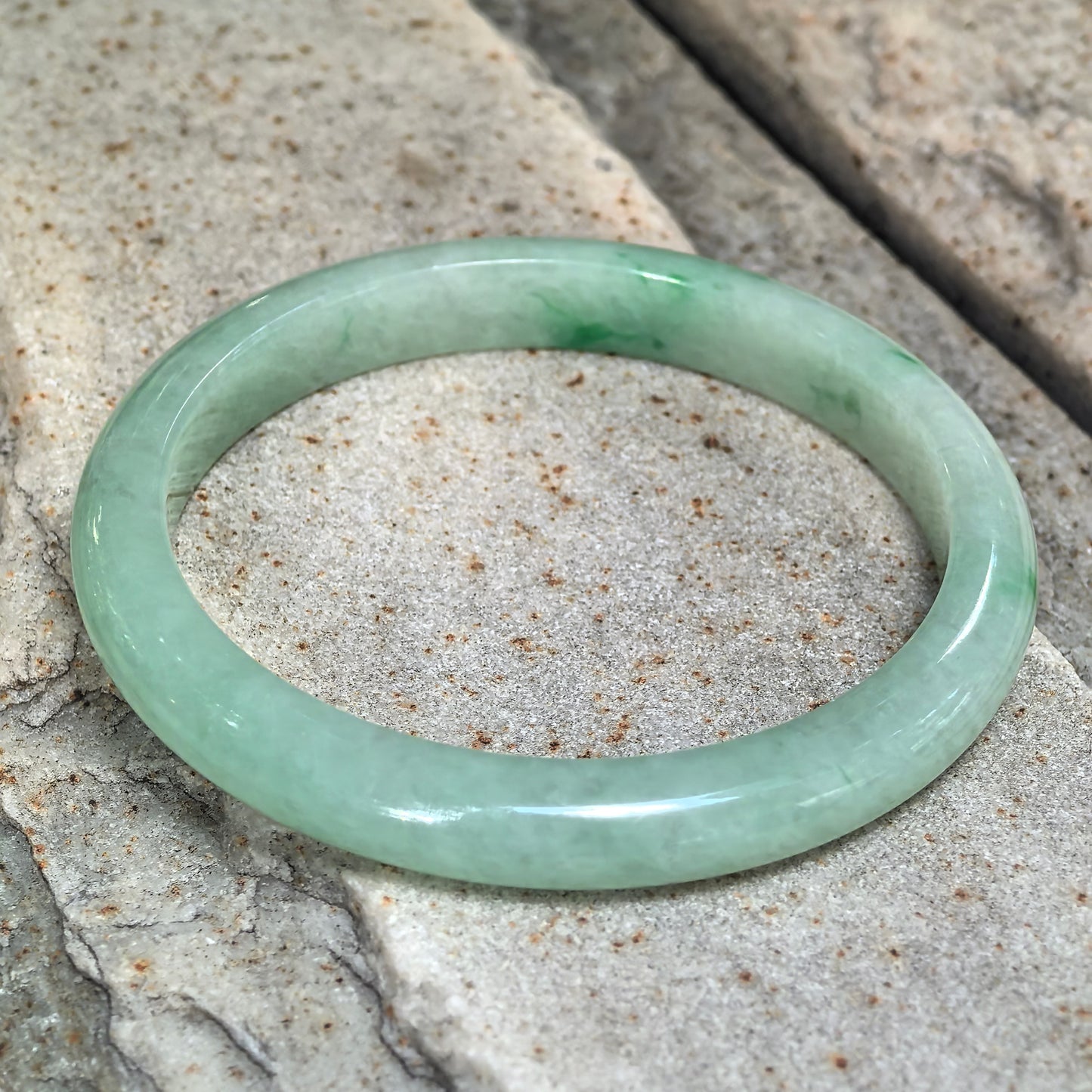 Apple green jadeite bangle resting at an angle on pale textured stone, with deeper green accents visible along the inner band.