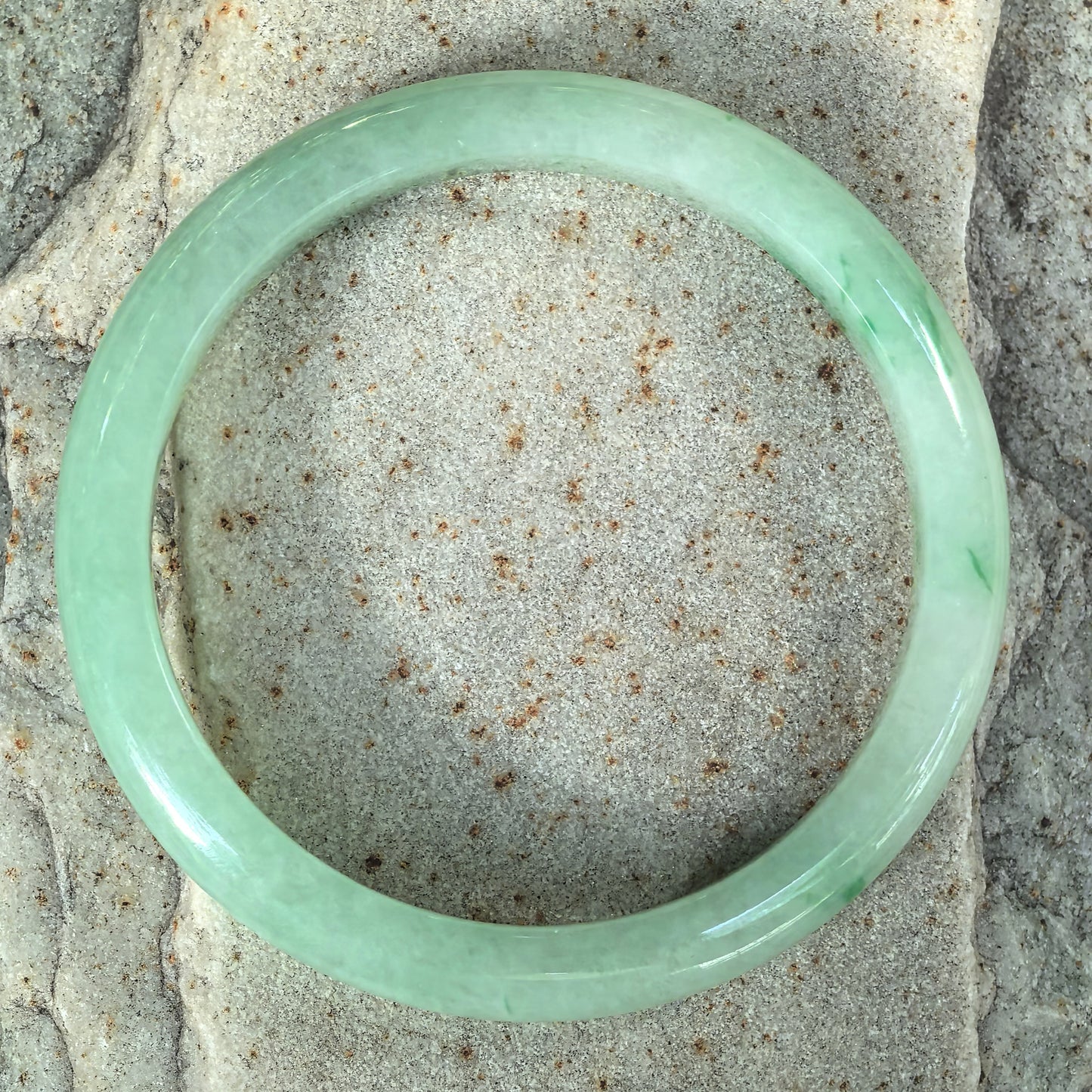 Top-down view of a slim green jadeite bangle on stone, showing color throughout the circular band and scattered darker green details.
