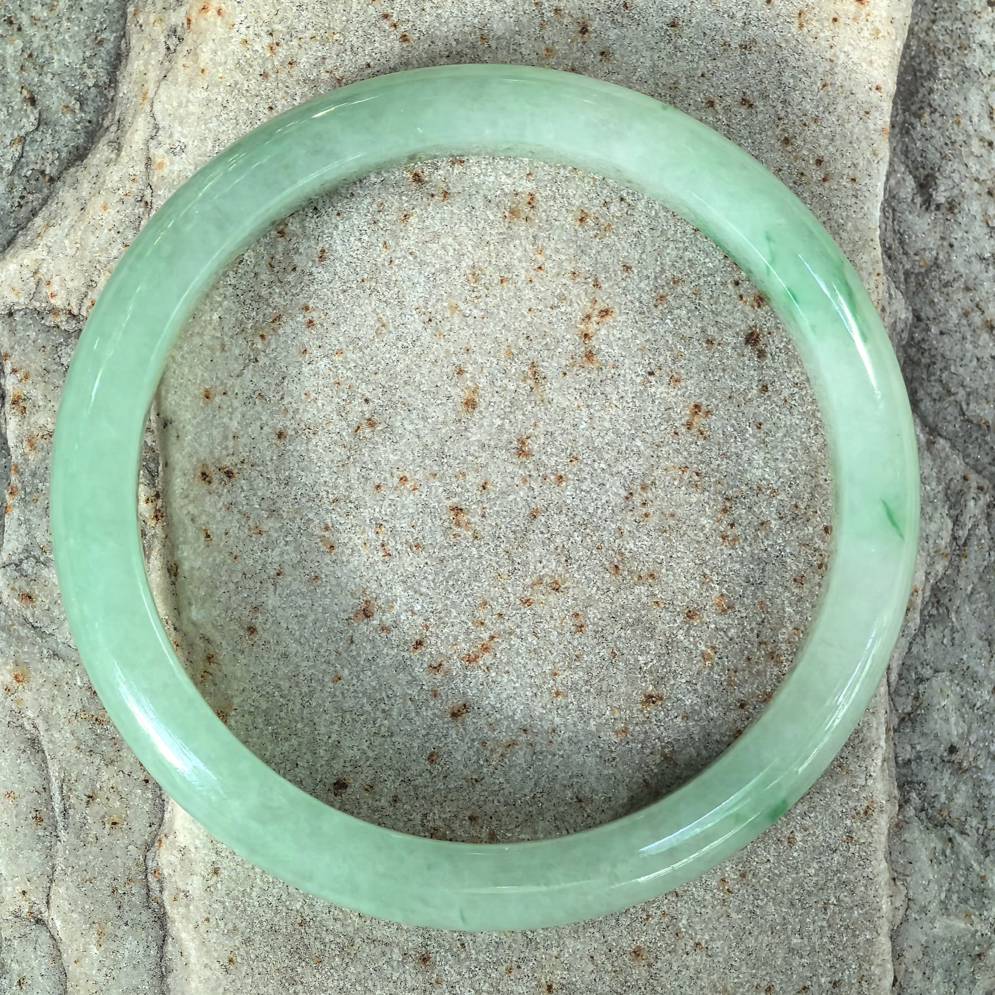 Top-down view of a slim green jadeite bangle on stone, showing color throughout the circular band and scattered darker green details.