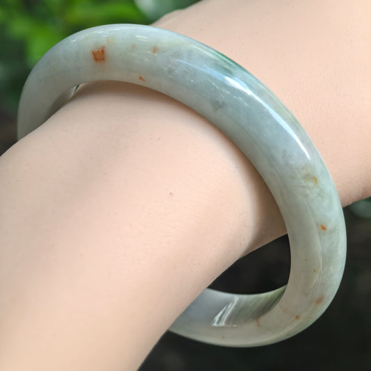 Angled wrist view of a pale green D-shape jadeite bangle with scattered natural golden-brown flecks.