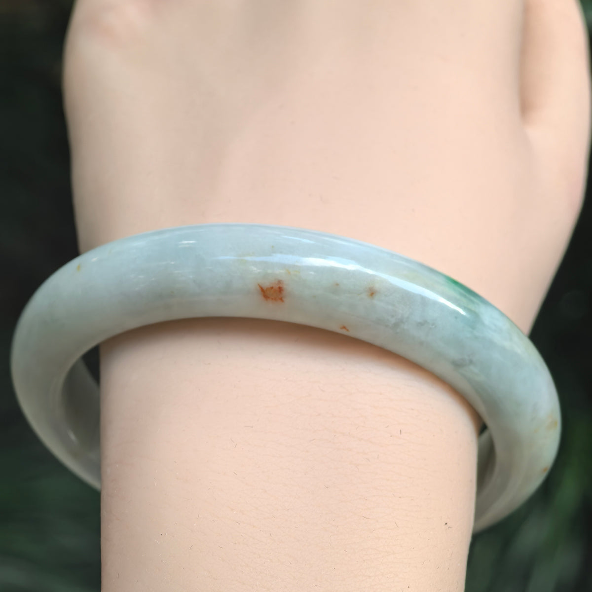 Front wrist view of a pale green jadeite bangle showing a prominent orange-brown marking and smaller warm-colored flecks.