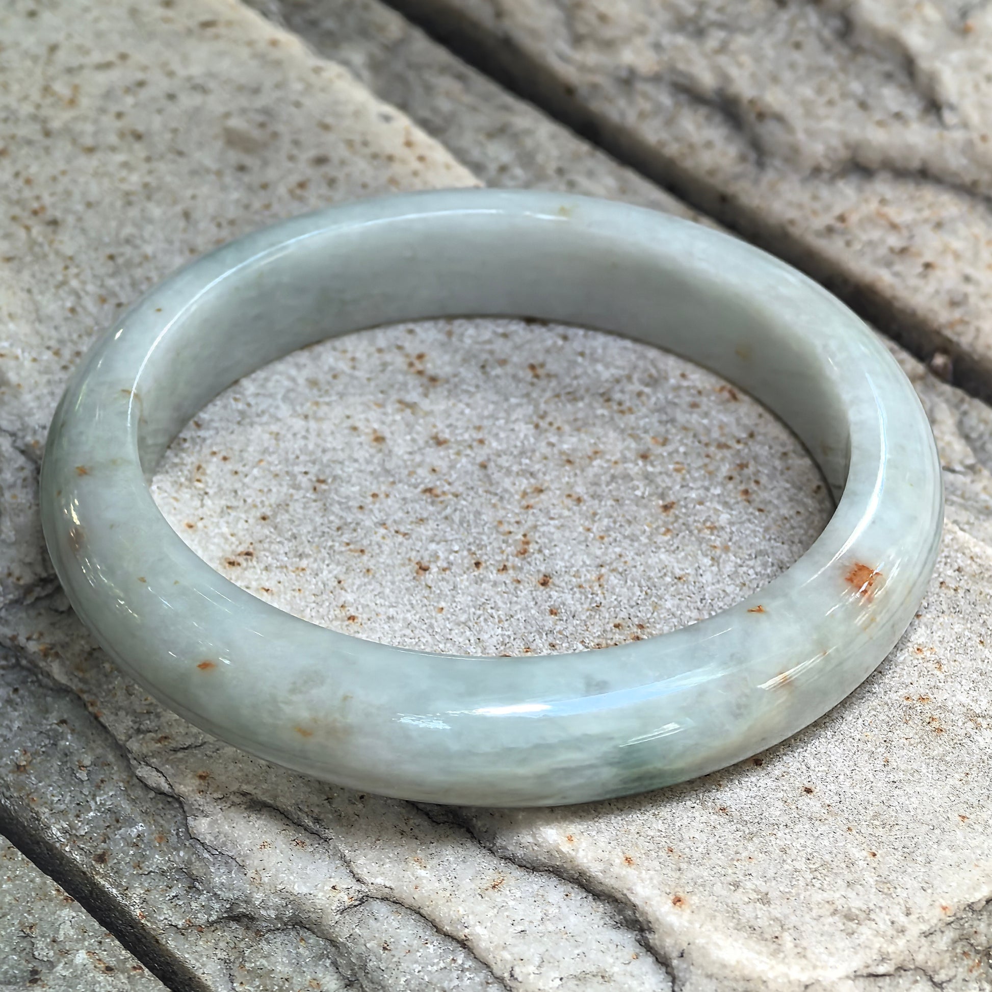 Pale green jadeite bangle resting at an angle on textured stone, showing its rounded exterior, flat interior and golden-brown accents.