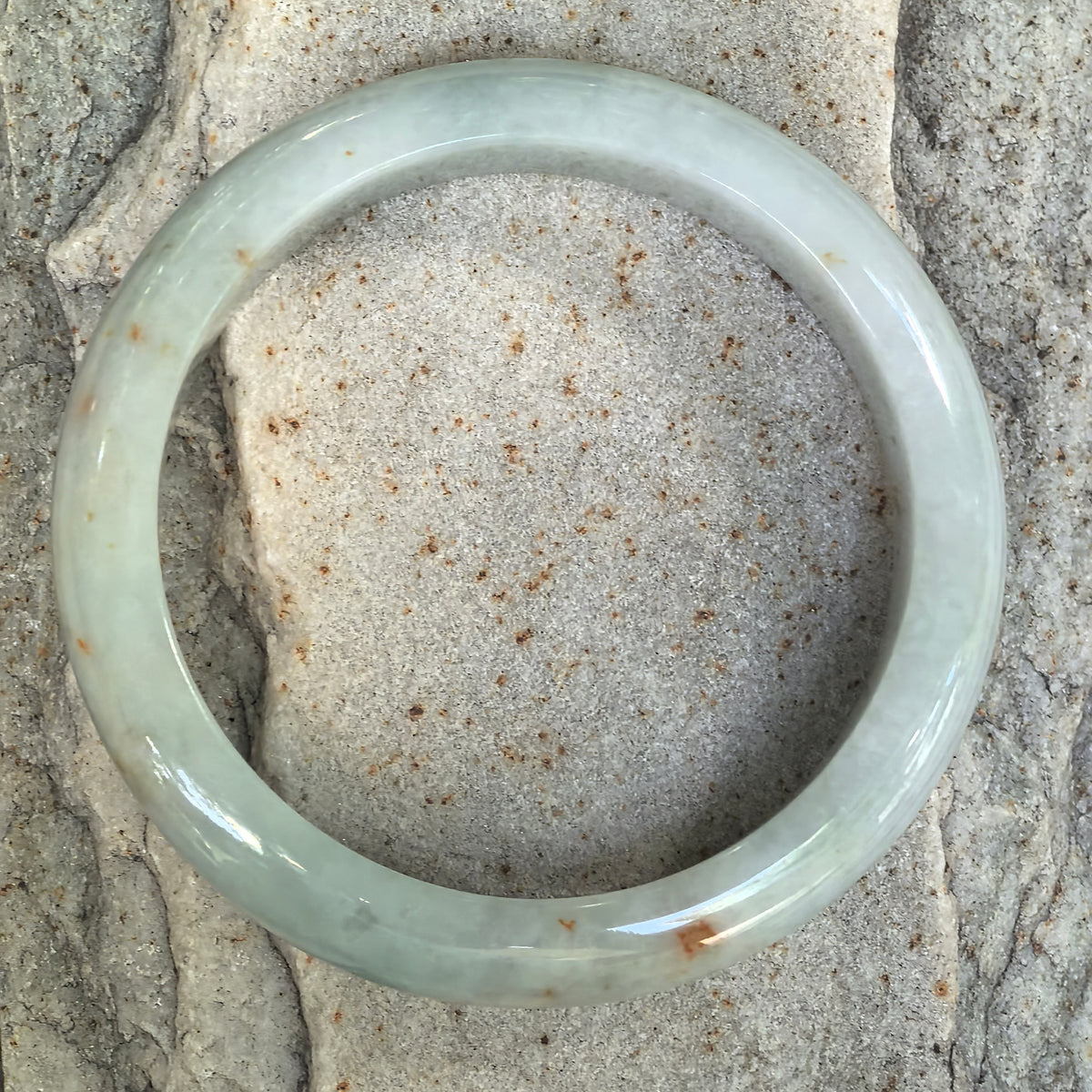 Top-down view of a pale green jadeite bangle on light stone, with scattered golden-brown flecks around the circular band.