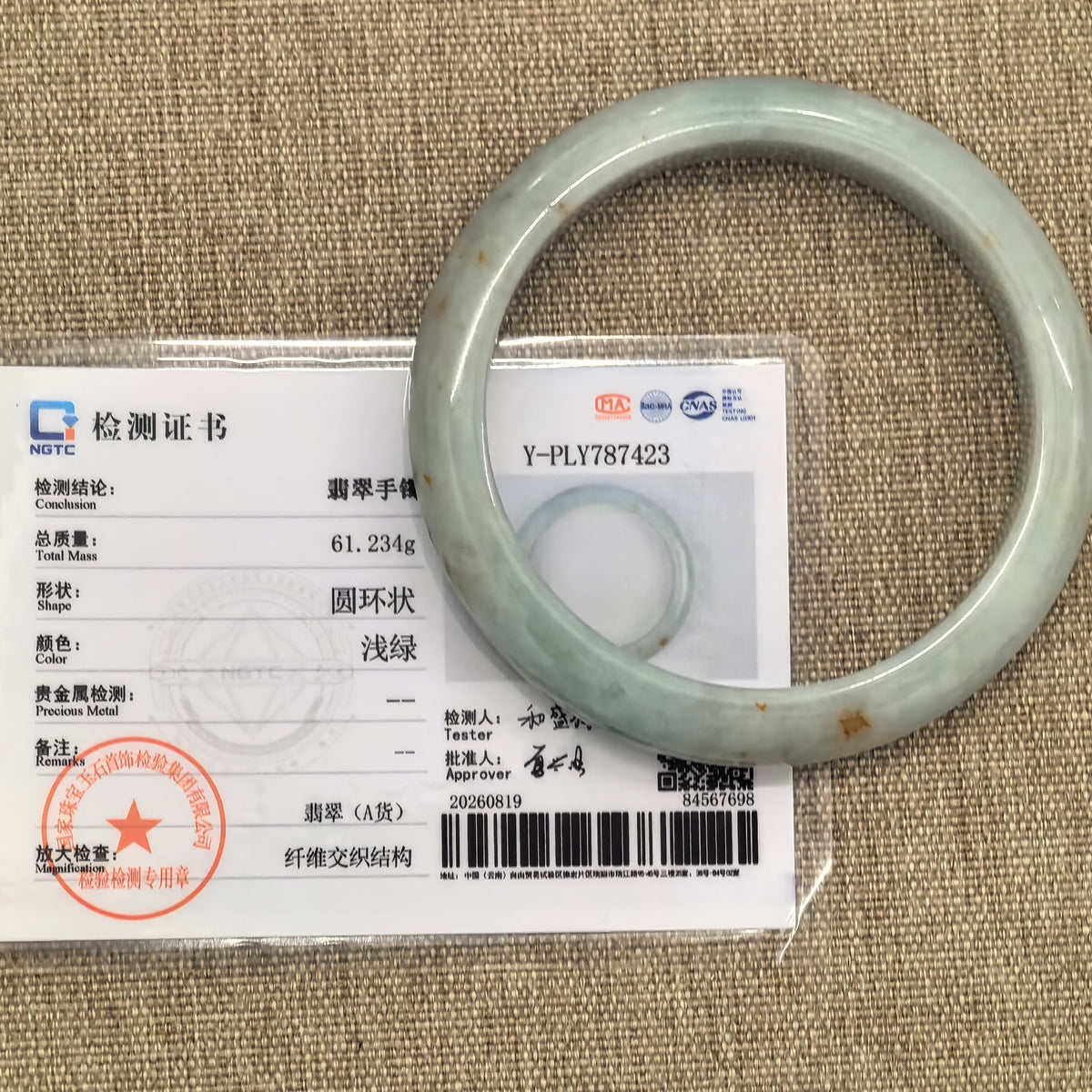 Pale green jadeite bangle with golden-brown flecks displayed beside its matching NGTC certificate on tan woven fabric.