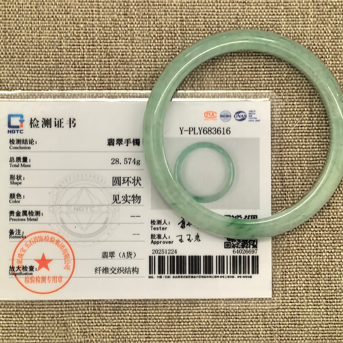 Apple green jadeite bangle displayed with its matching NGTC certificate on tan woven fabric.
