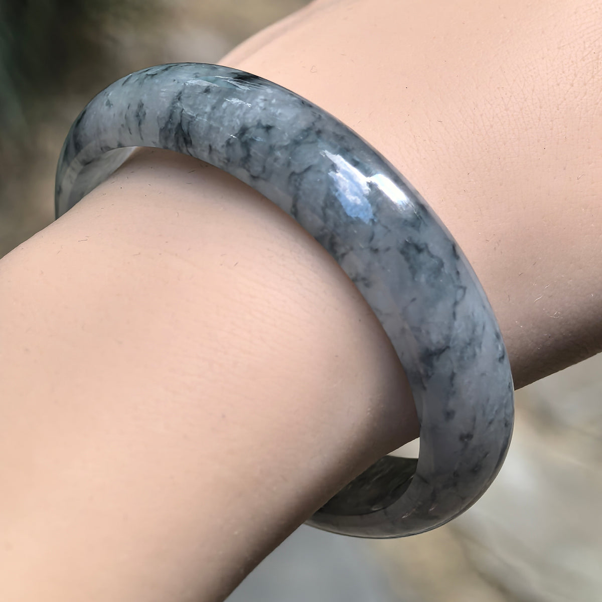 Angled wrist view of an oval jadeite bangle with inky blue floating patterns across a pale gray base.