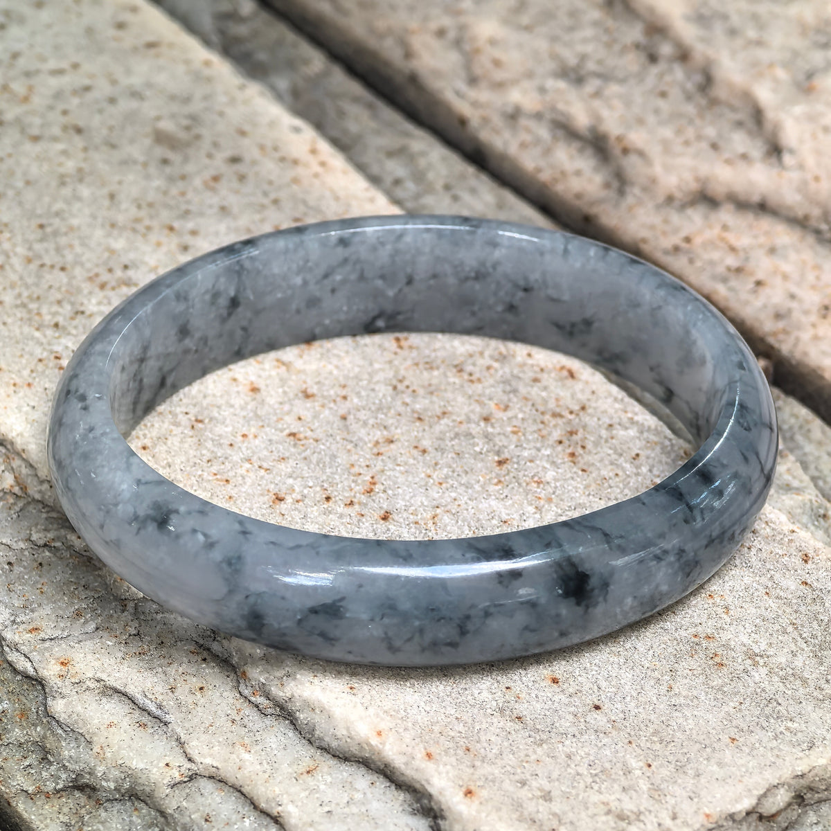Oval jadeite bangle resting at an angle on pale stone, showing its inner surface and scattered blue floating patterns.