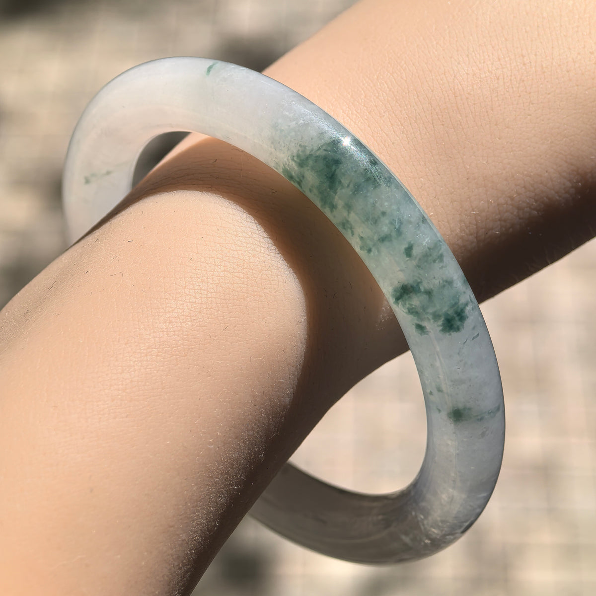 Sunlit wrist close-up of a round-bar jadeite bangle, highlighting its translucent pale base and green floating patterns.