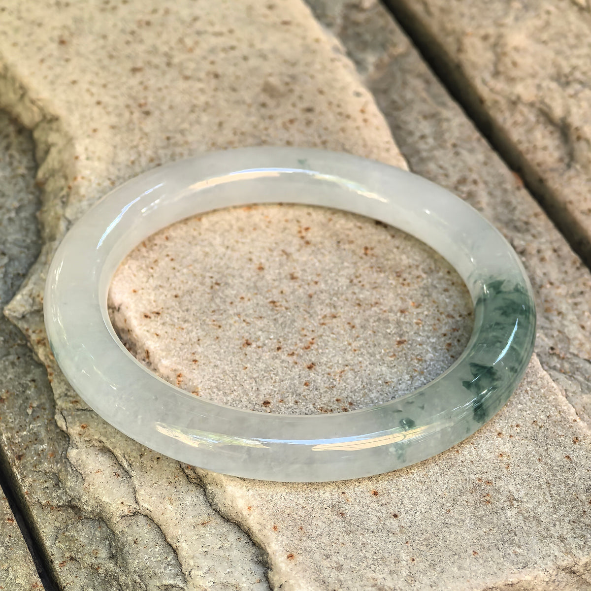 Angled view of a translucent white jadeite bangle resting on pale stone, with green floating patterns concentrated along the right side.