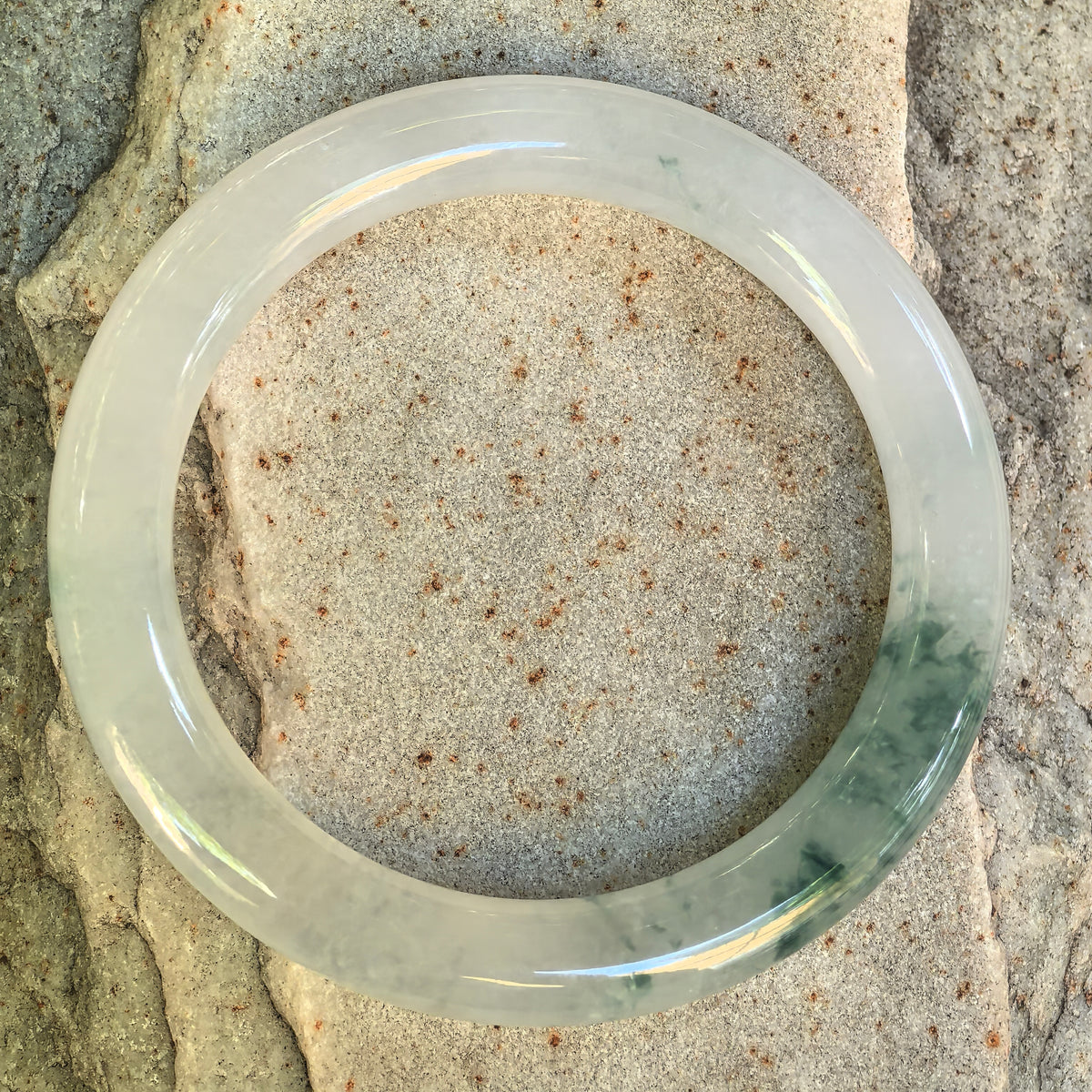 Top view of a round-bar jadeite bangle on textured stone, showing its circular form, pale base and scattered green markings.