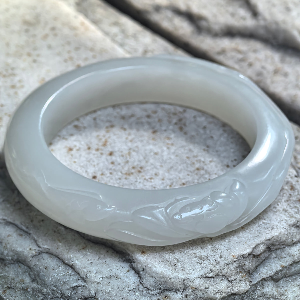 Angled view of a white Hetian jade D-shape bangle on stone, showing lotus carving around the rounded exterior and its flatter inner face.