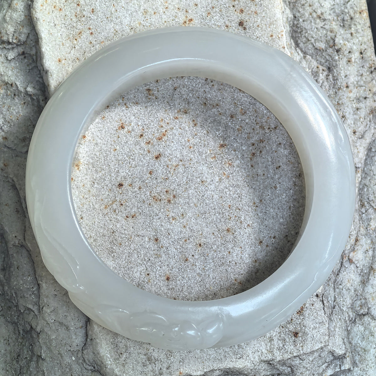 Top view of a white nephrite lotus-carved D-shape bangle resting on stone, showing its circular outline and subtle celadon undertone.