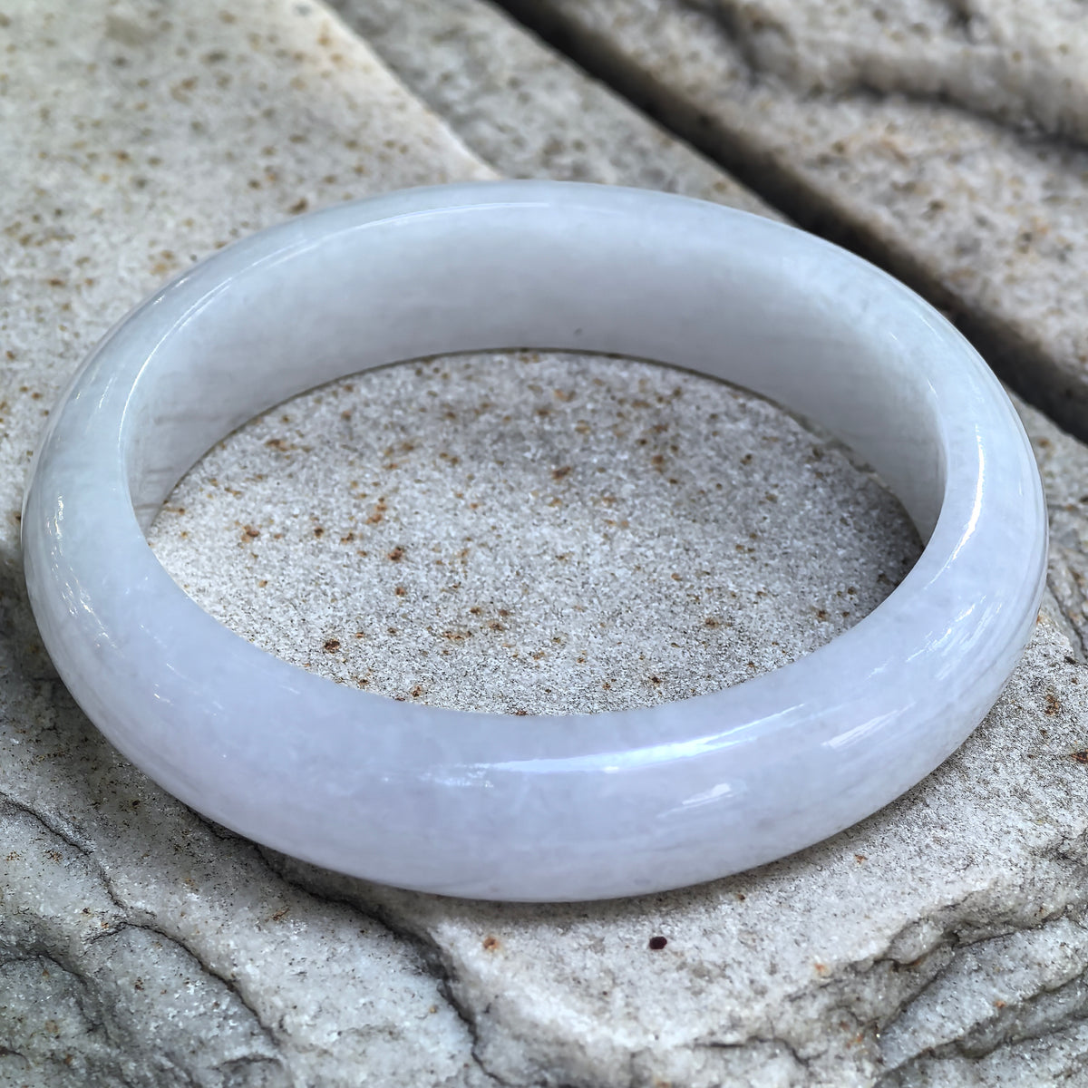 White jadeite D-shape bangle resting at an angle on textured stone, showing its rounded exterior and flat inner surface.