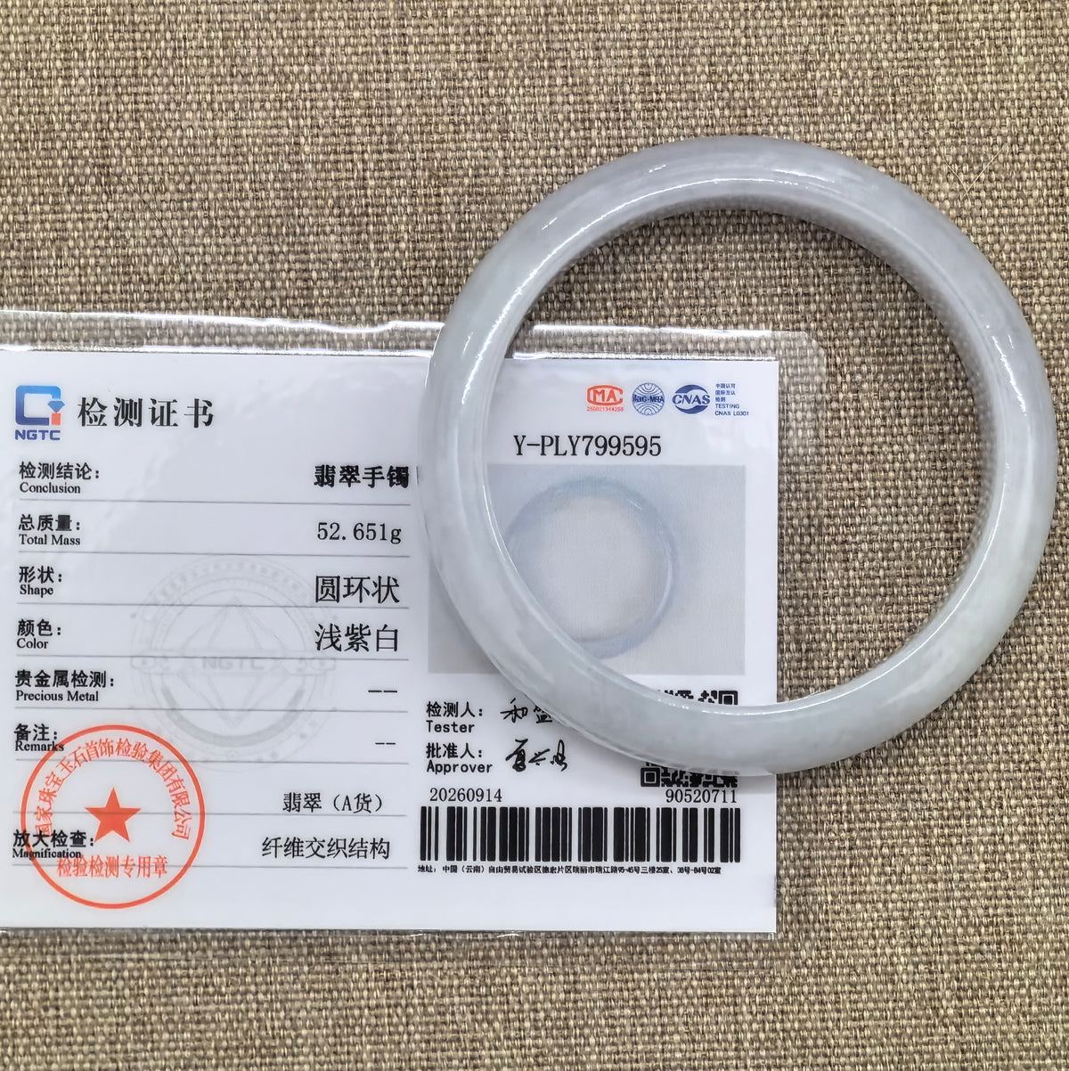 White jadeite bangle displayed with its matching NGTC certificate on a woven fabric background.