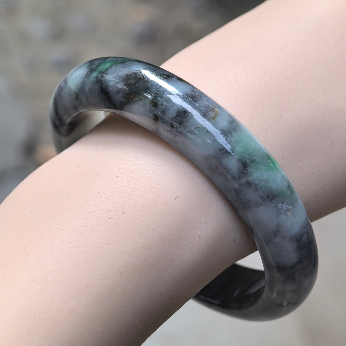 Angled wrist view of a glutinous jadeite bangle showing smoky black patterns, gray areas, and localized green accents.