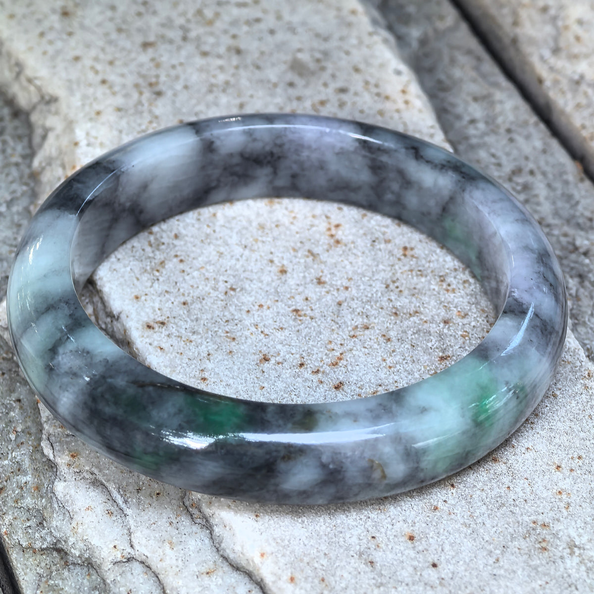 Gray and black D-shape jadeite bangle resting at an angle on pale stone, showing its flat inner face and contrasting natural colors.