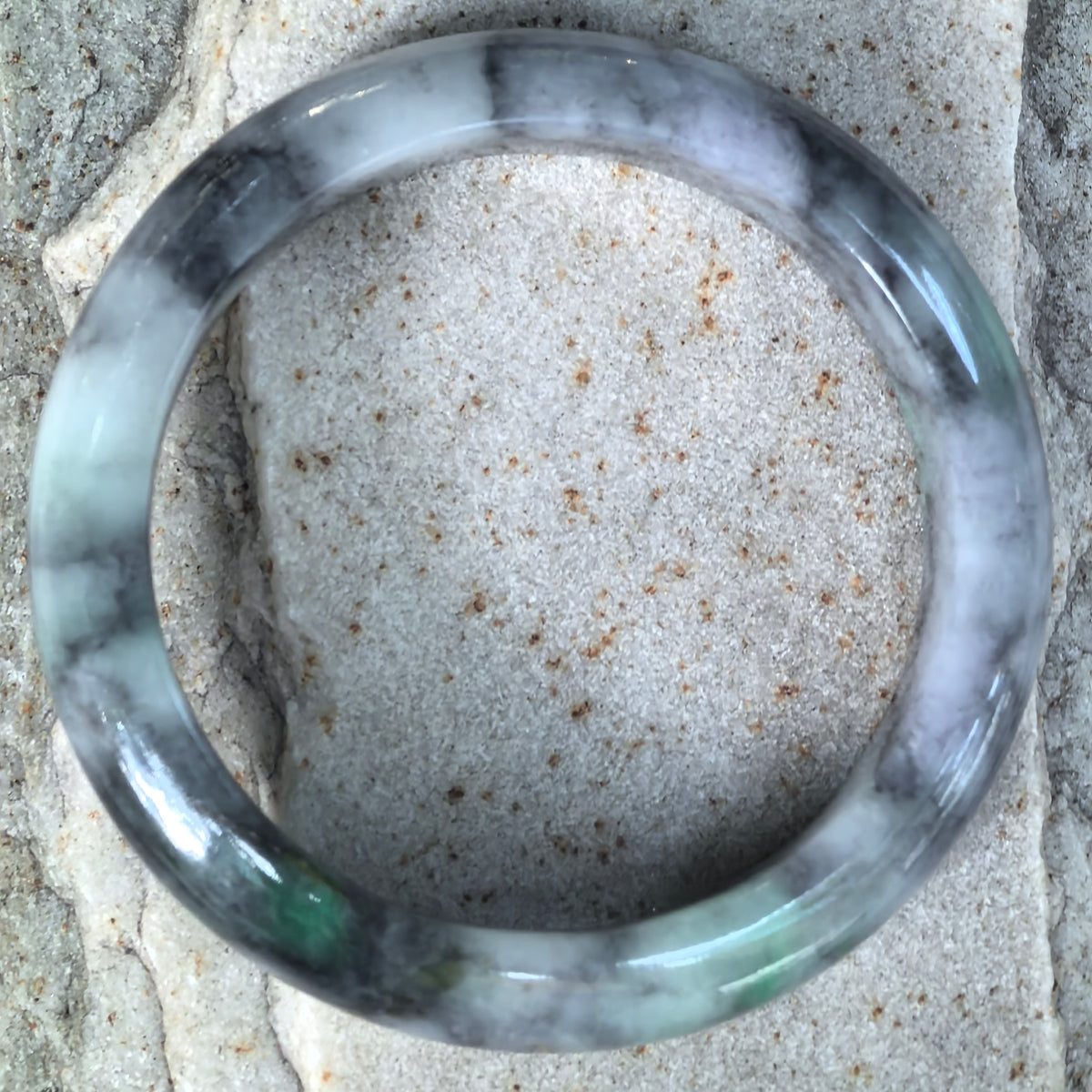 Top view of a gray and black jadeite bangle on pale stone, showing its circular outline and green accents along the lower section.