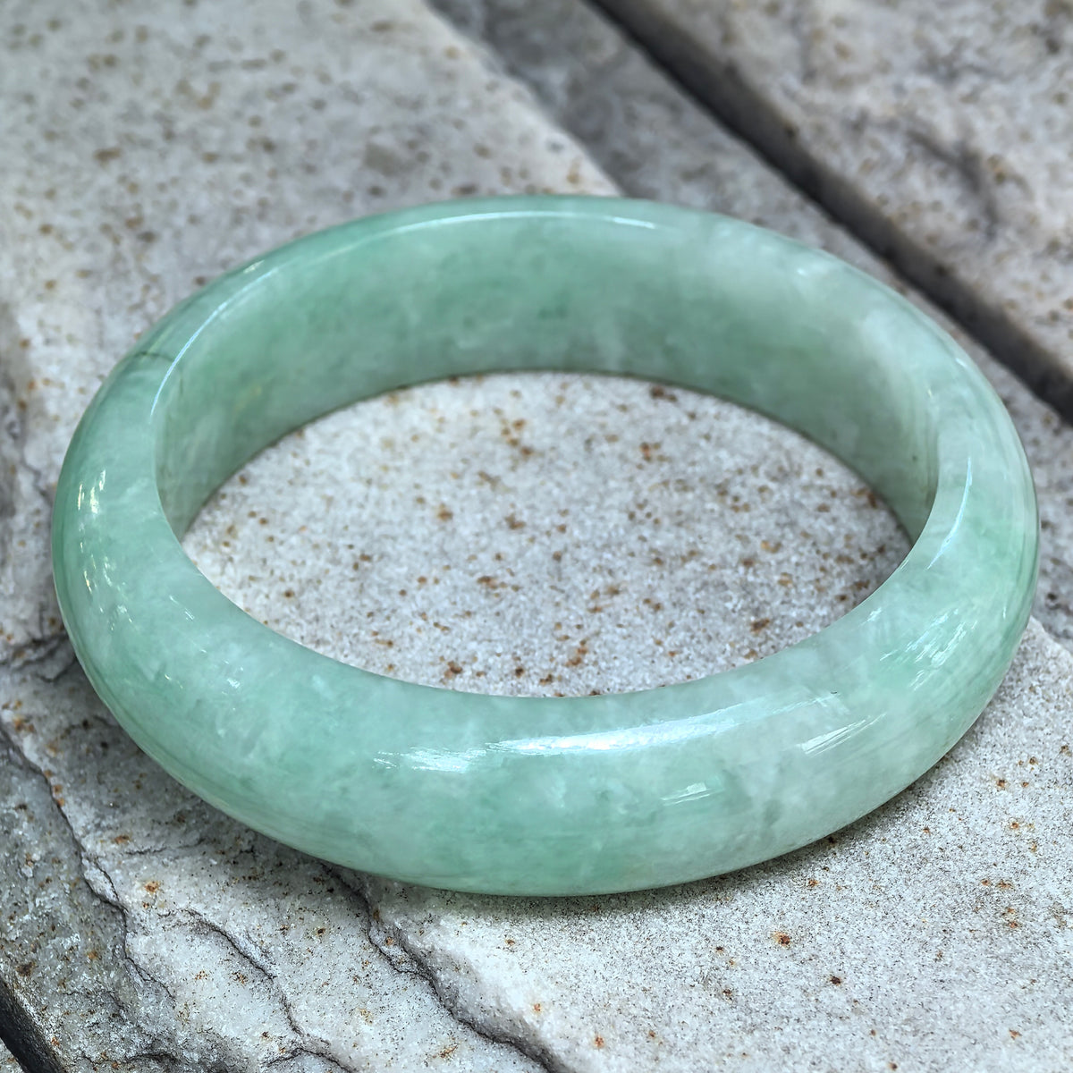 Apple green D-shape jadeite bangle resting at an angle on pale stone, showing the broad band and flat inner face.