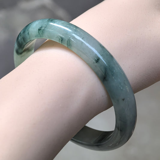Angled wrist view of a jadeite D-shape bangle with deep green floating patterns across a light green base and a polished rounded exterior.